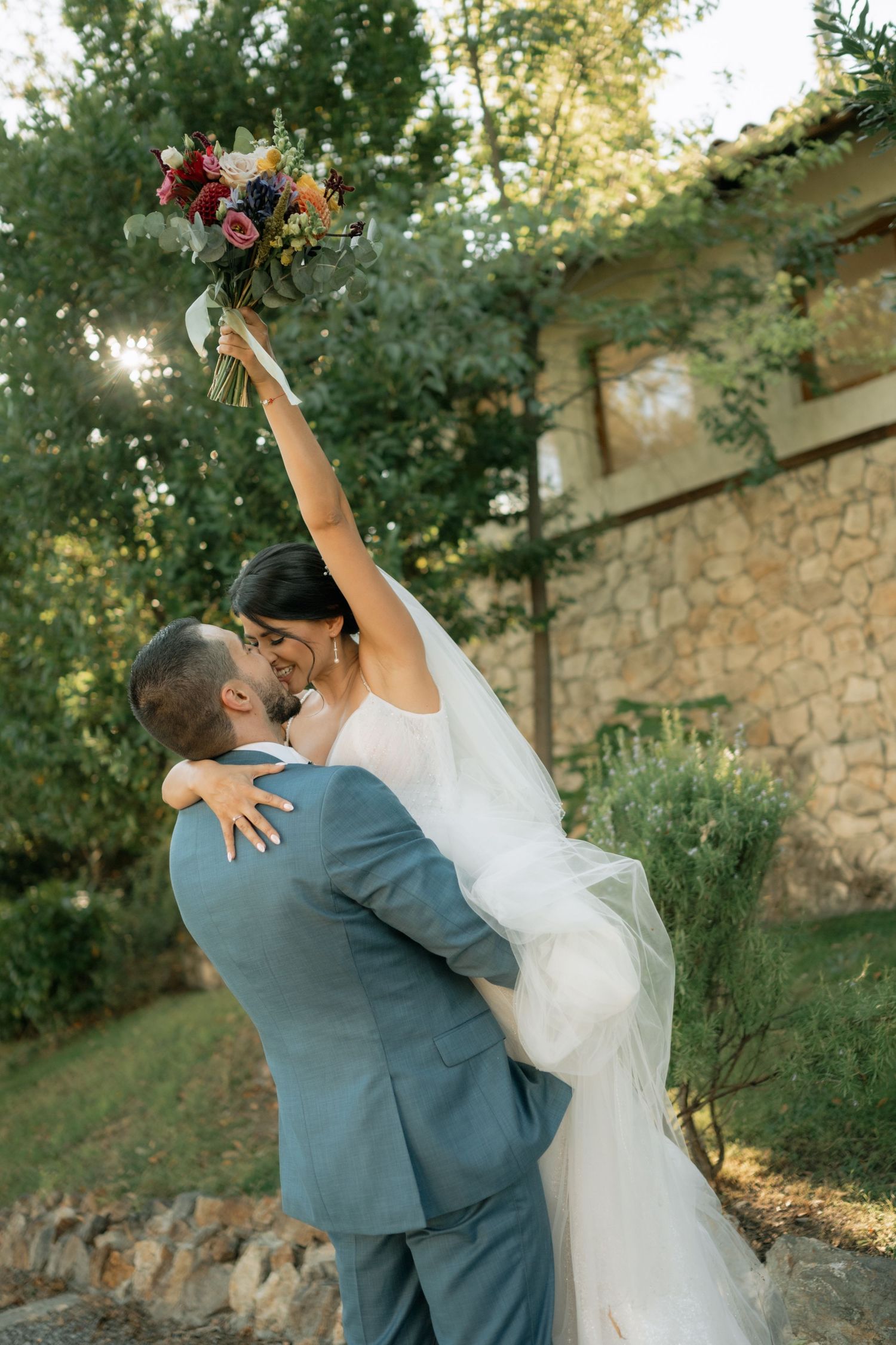 A joyful wedding couple embraces and spins together outdoors near a stone wall during golden hour.