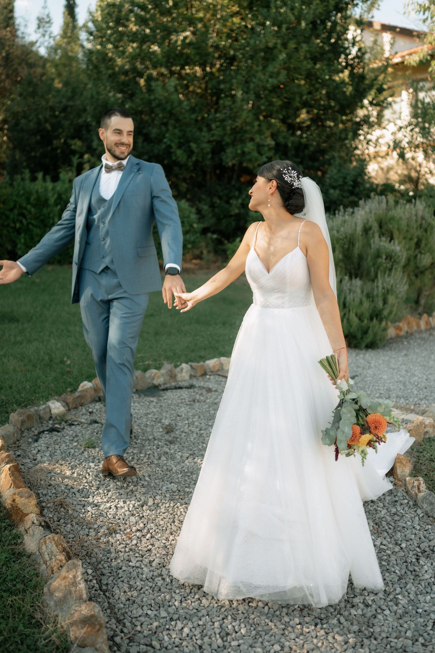 A couple in wedding attire walks along a gravel path in a garden setting during their special day.