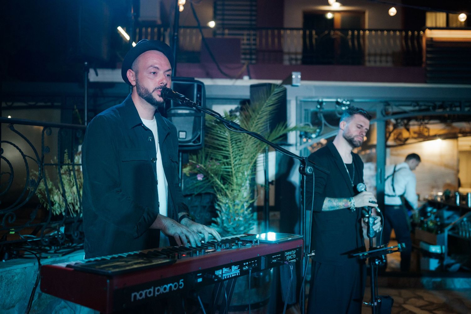 Musicians perform on stage with electronic equipment and instruments in a dimly lit venue setting.