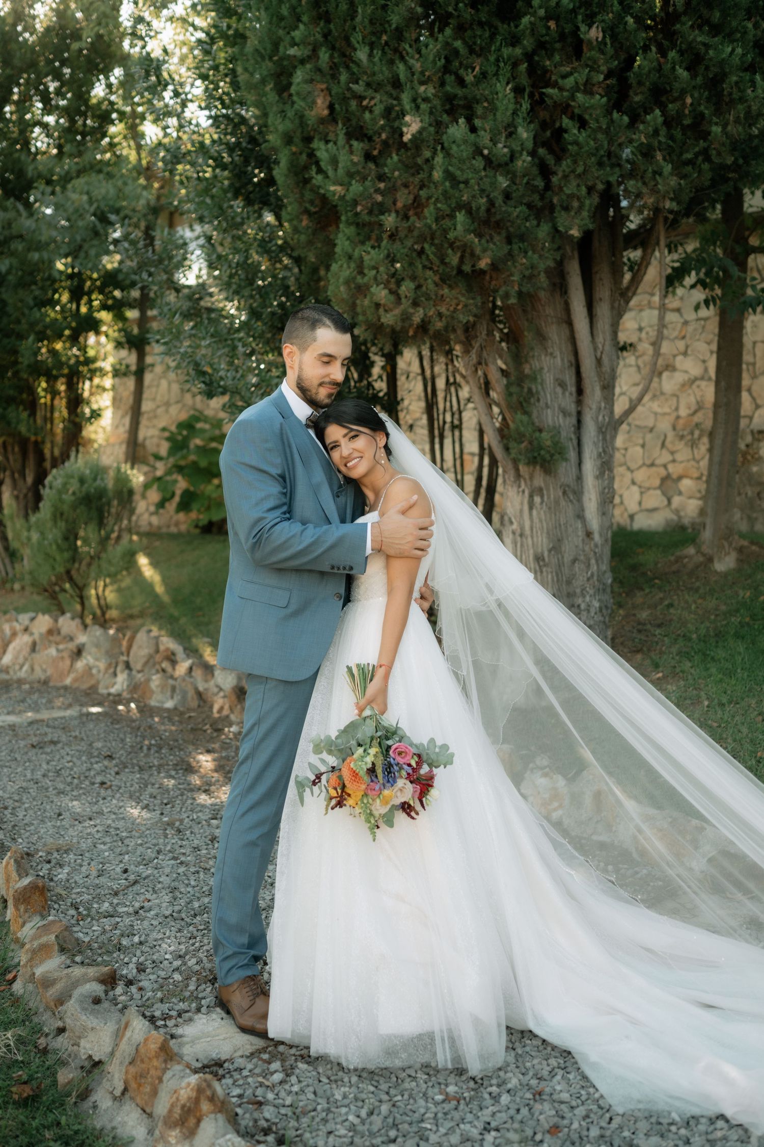 A romantic wedding couple embraces outdoors with a flowing white veil and floral bouquet in a wooded setting.