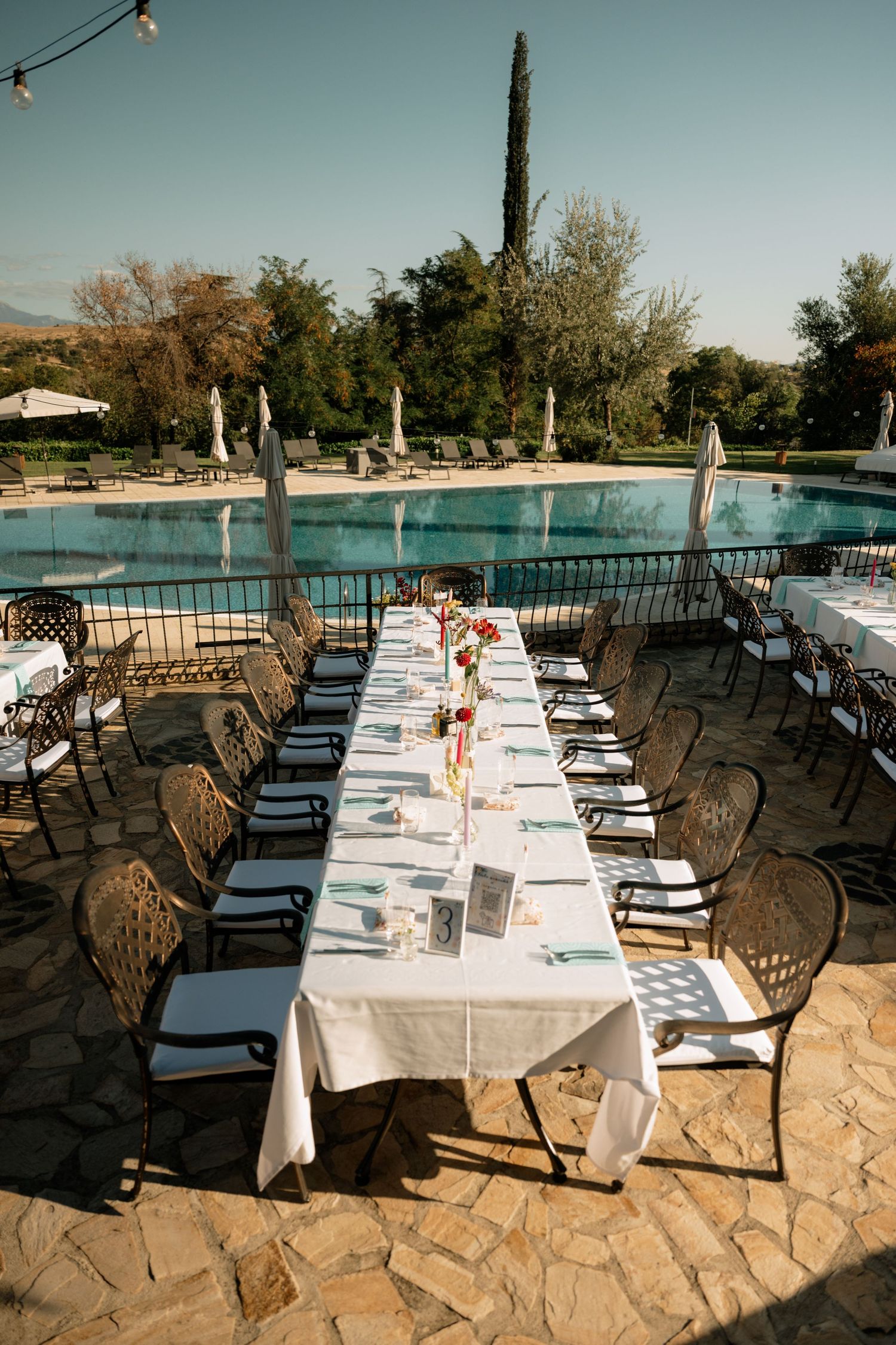 Elegant outdoor dining setup with long white-clothed tables and ornate metal chairs on a stone patio overlooking a pool.