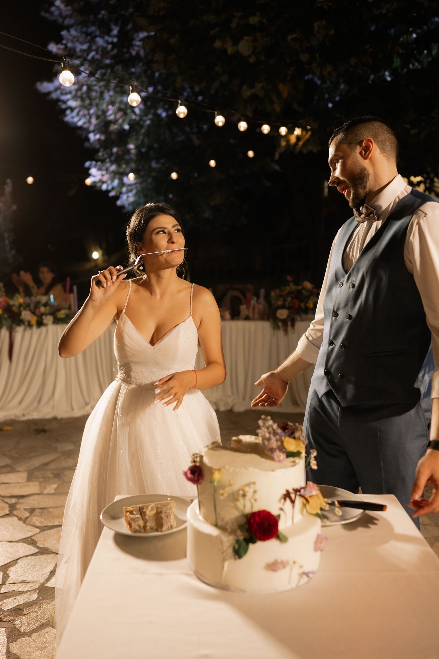 A couple shares a moment during their wedding reception as they taste cake at a table decorated with string lights outdoors.