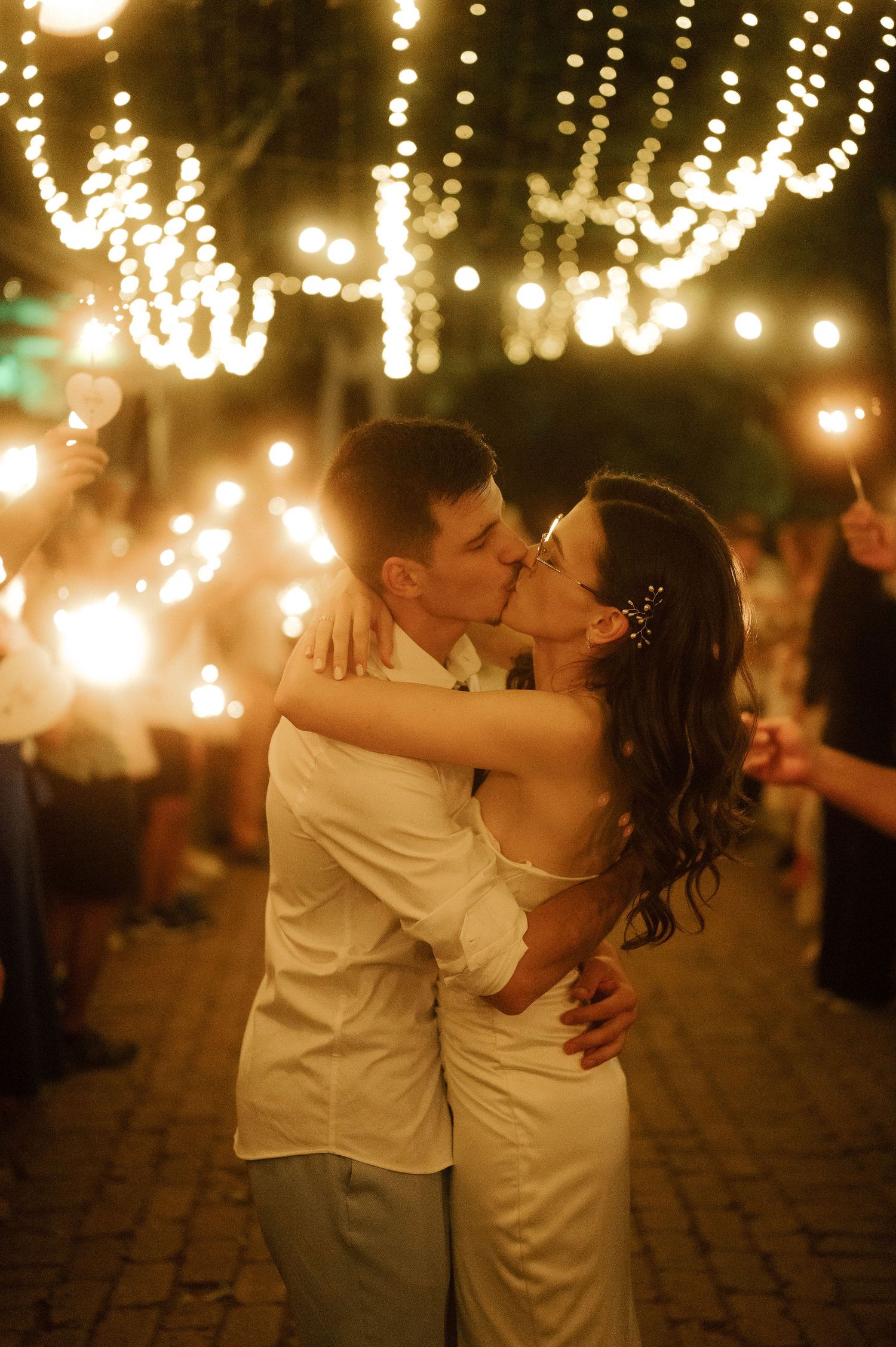 A romantic couple shares a kiss under twinkling string lights at an outdoor evening celebration.
