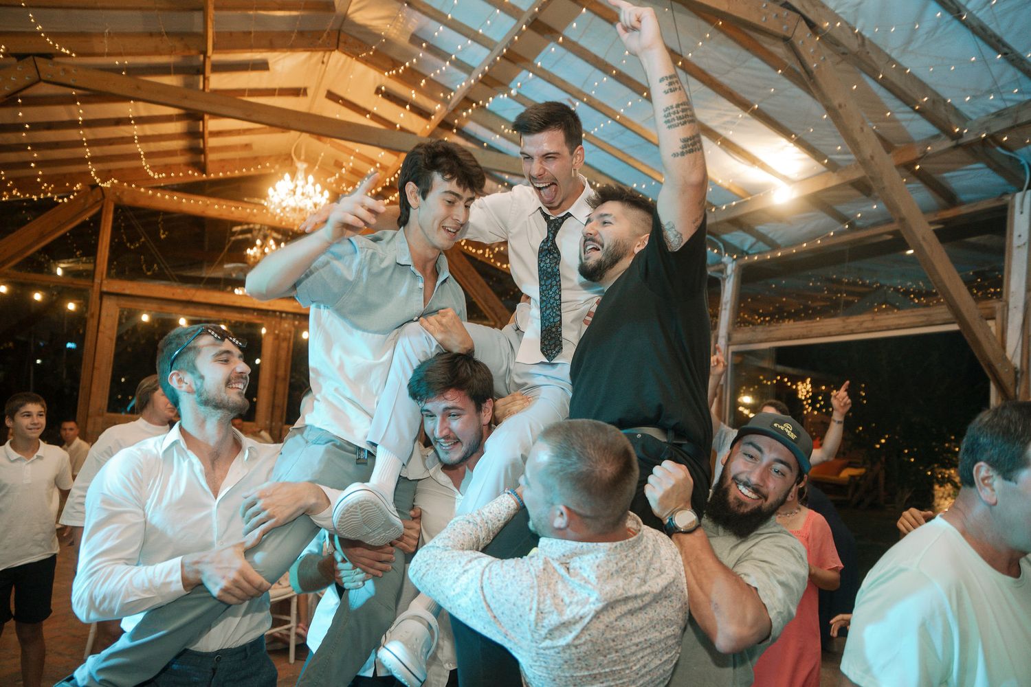 A group of people dancing and celebrating at a lively party under wooden beams with warm lighting.