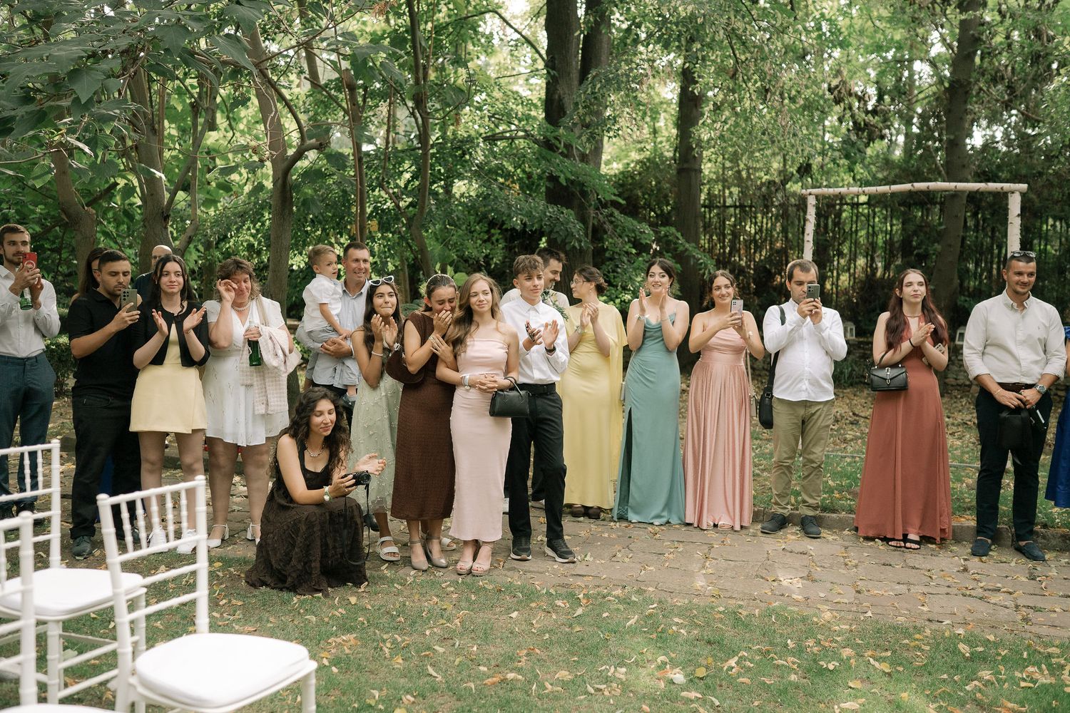 Wedding guests in formal attire gather outdoors under trees for a ceremony with white chairs and an arch.