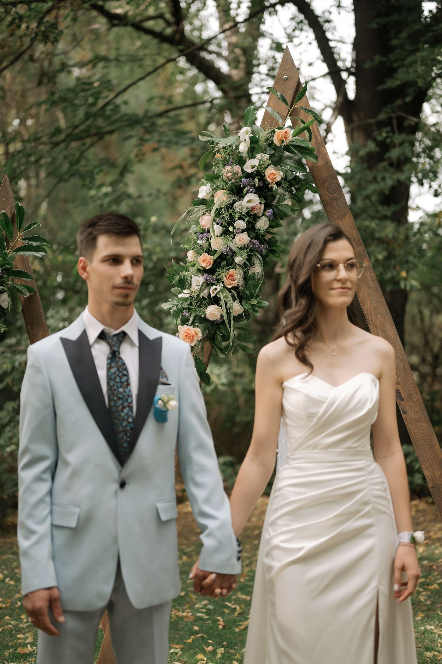 A wedding couple poses in a wooded setting with floral arch decorations and natural greenery.