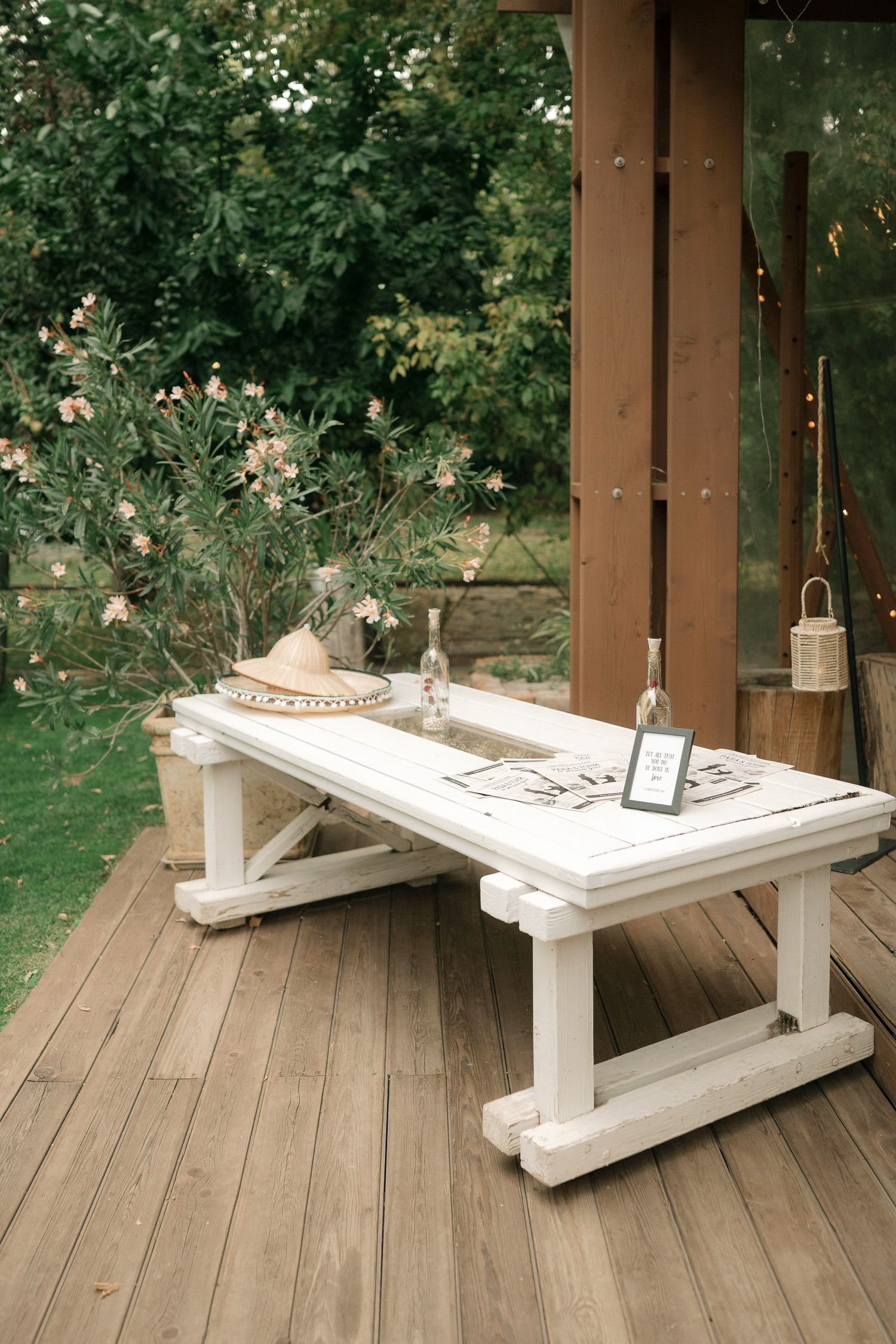 A rustic white wooden bench table sits on a deck with decorative items and flowering bushes in the background.