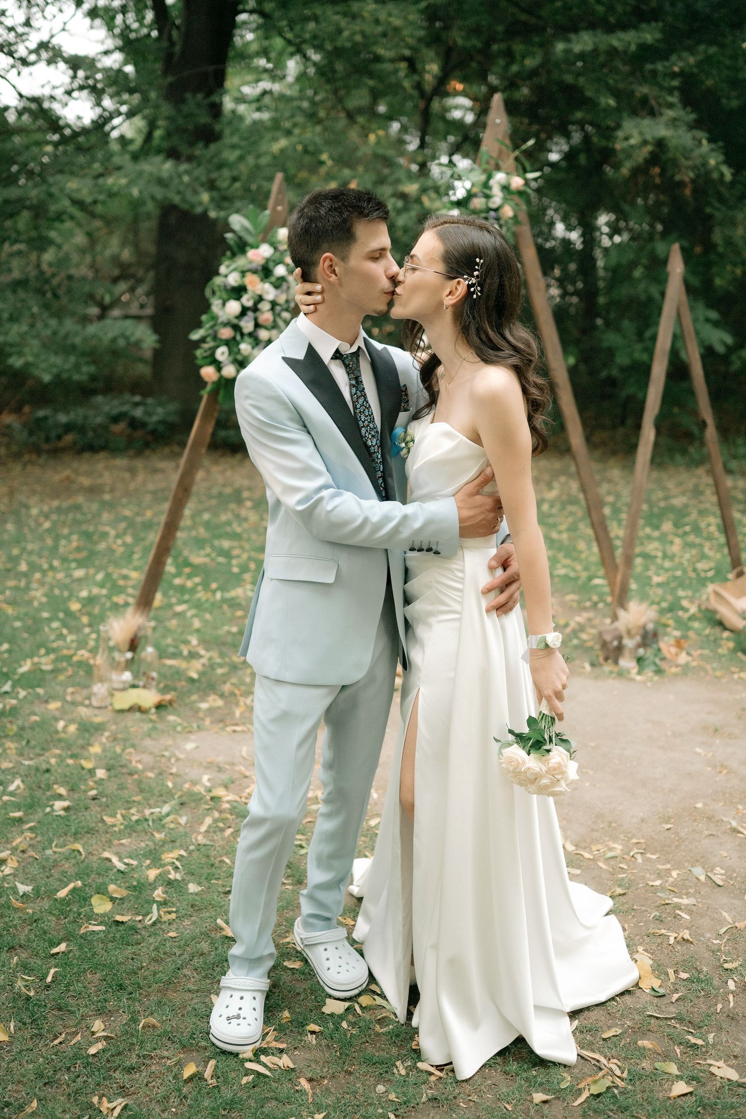Newlywed couple shares romantic moments in a wooded outdoor setting wearing white wedding attire.