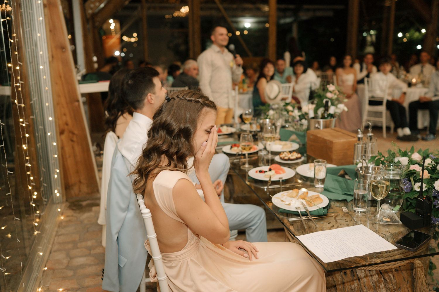 Wedding guests enjoying dinner at rustic wooden tables decorated with string lights in a barn venue.
