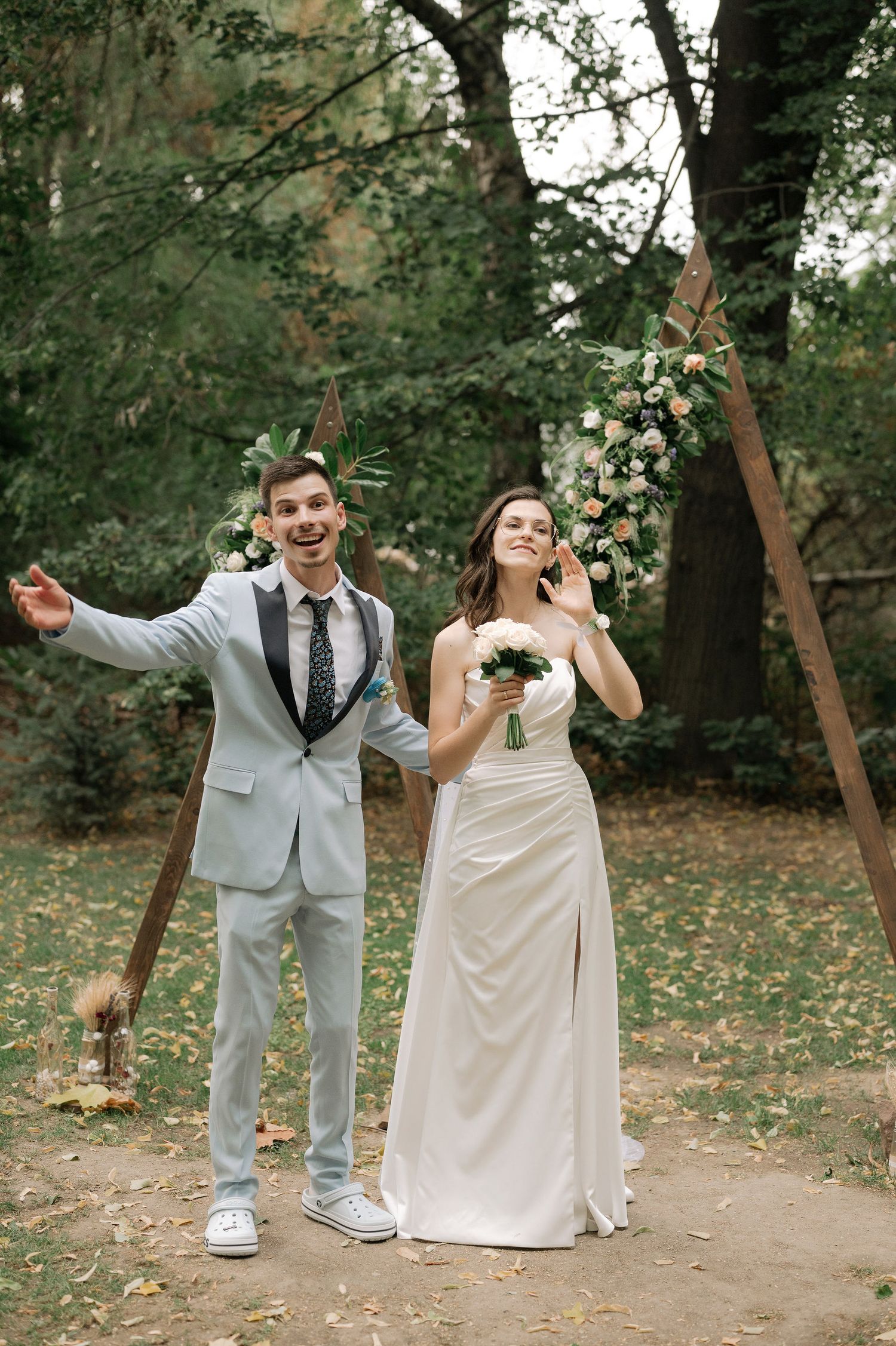 A couple in wedding attire poses playfully on a forest path during an autumn outdoor ceremony.