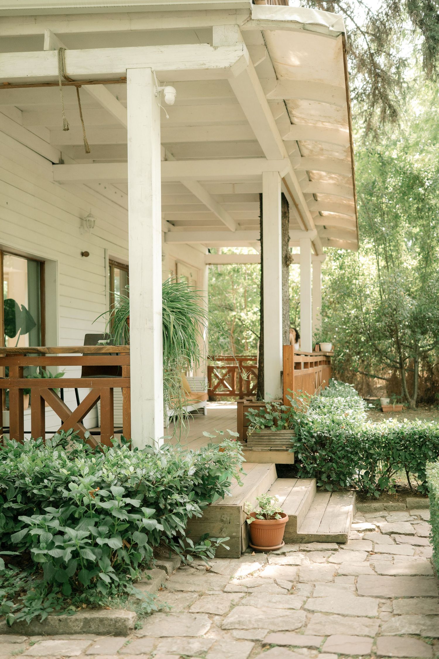 Serene covered porch with white columns and stone flooring surrounded by lush greenery and potted plants.
