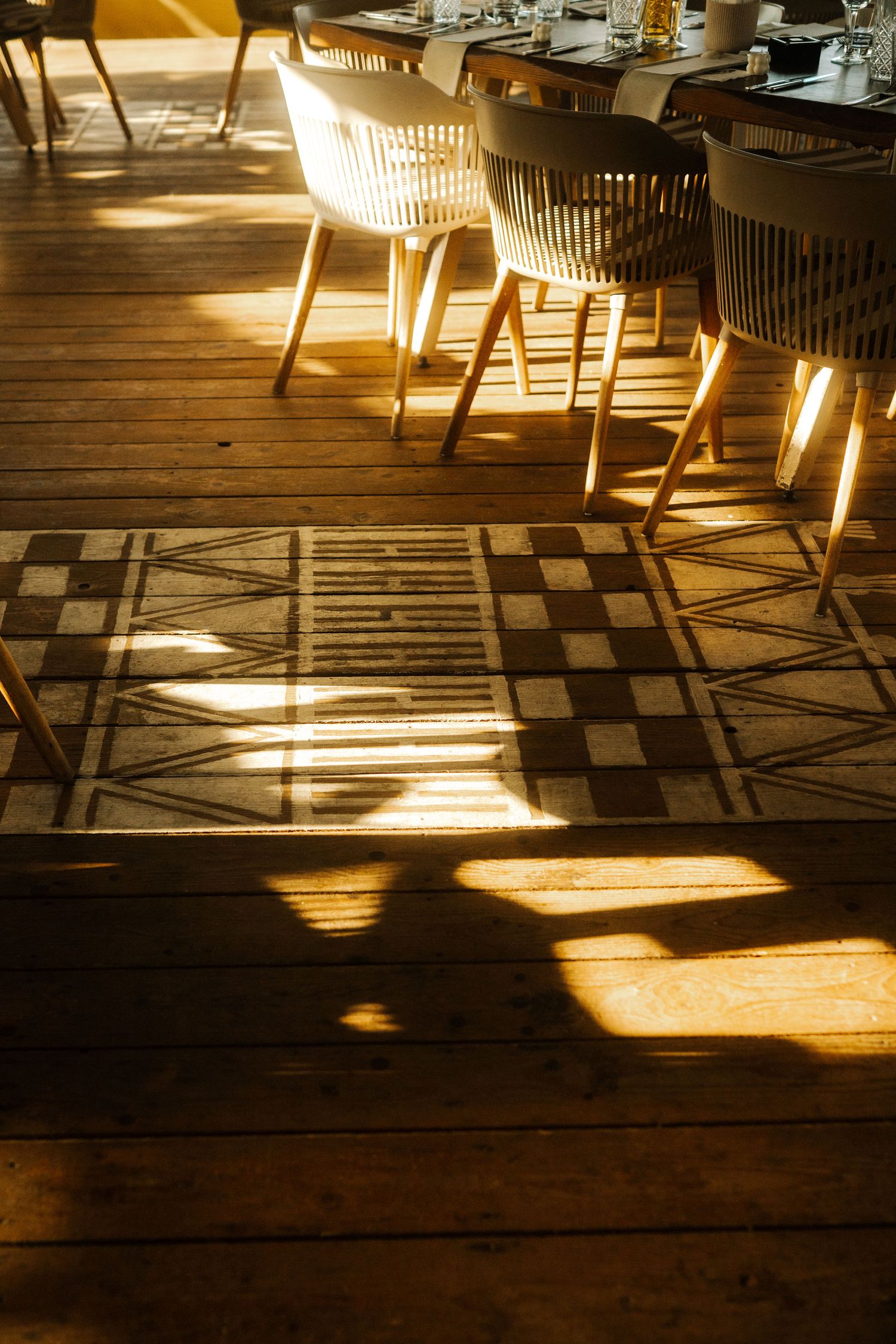 Sunlight casts dramatic shadows of wooden chairs on a checkered wooden floor in a restaurant interior.