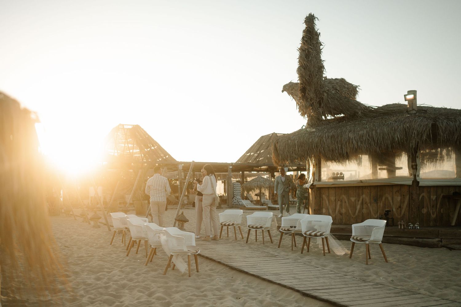 A row of beach chairs and umbrellas line a sandy shoreline at sunset with golden light filtering through the scene.
