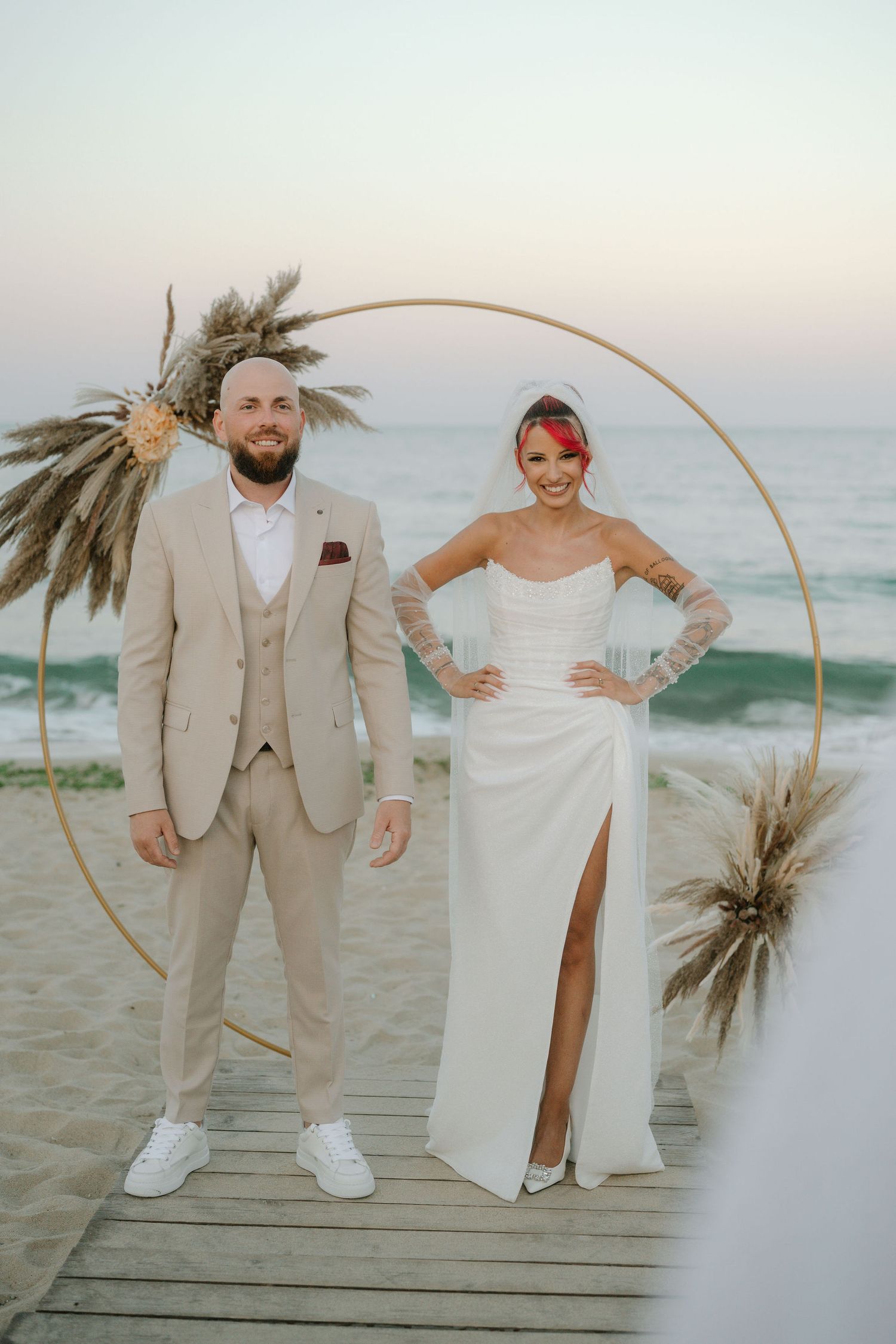 A couple poses on a beach boardwalk at sunset with dried pampas grass decor and a circular arch backdrop.