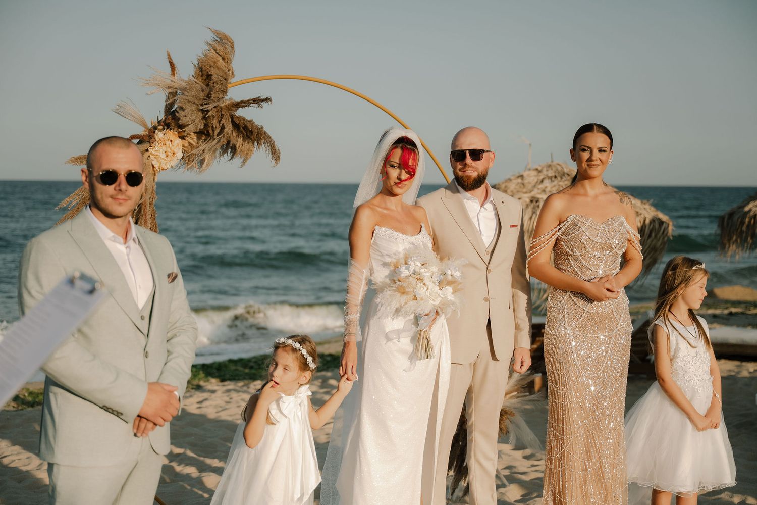 Wedding party poses together on a beach at sunset with palm trees and ocean waves in the background.
