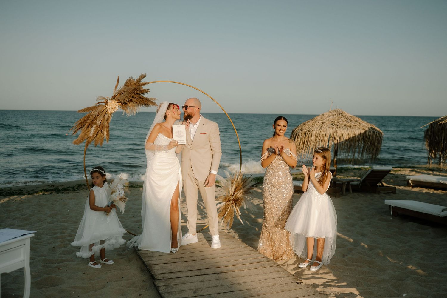 A beachside wedding party in white attire walks along a wooden pier at sunset with flowing palm fronds in the breeze.
