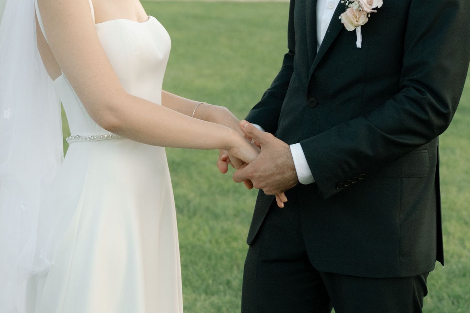 A bride and groom hold hands during an outdoor wedding ceremony on a green lawn.