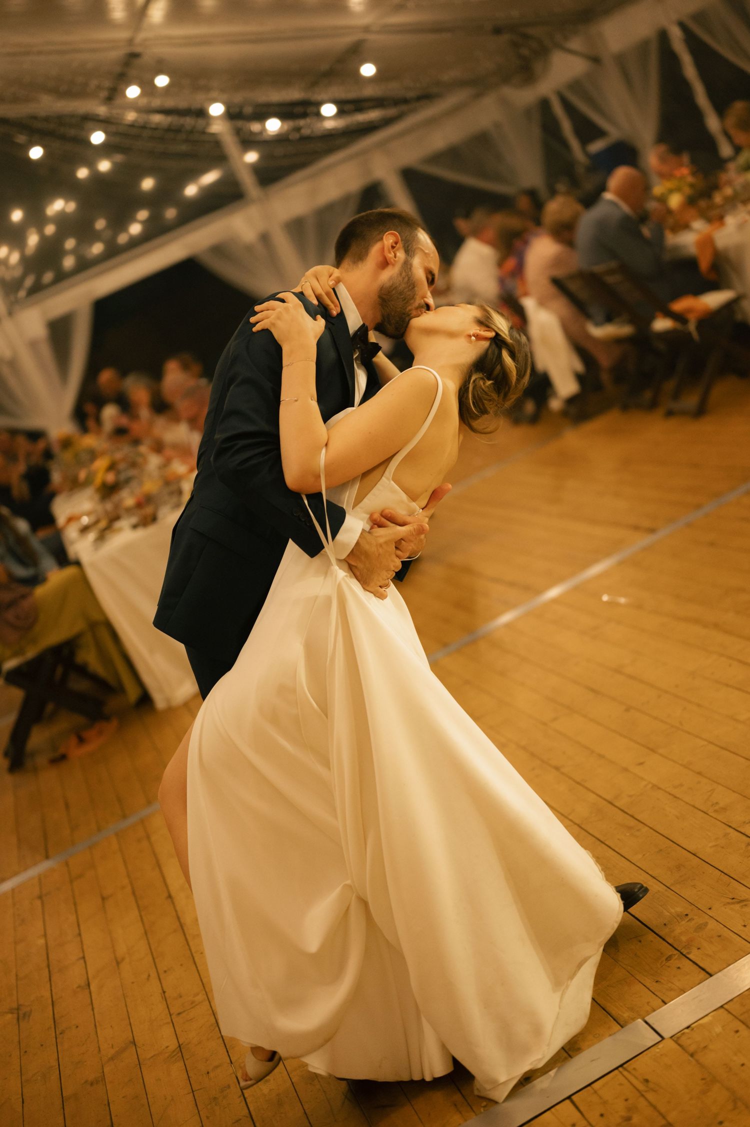 A couple performs a romantic dip dance move on a wooden dance floor under twinkling string lights at their wedding reception.