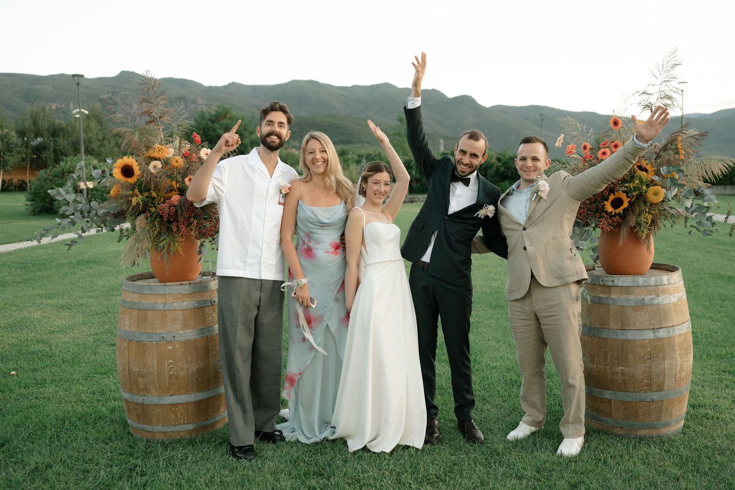Wedding party poses outdoors in front of rustic wine barrels with mountain scenery in the background.