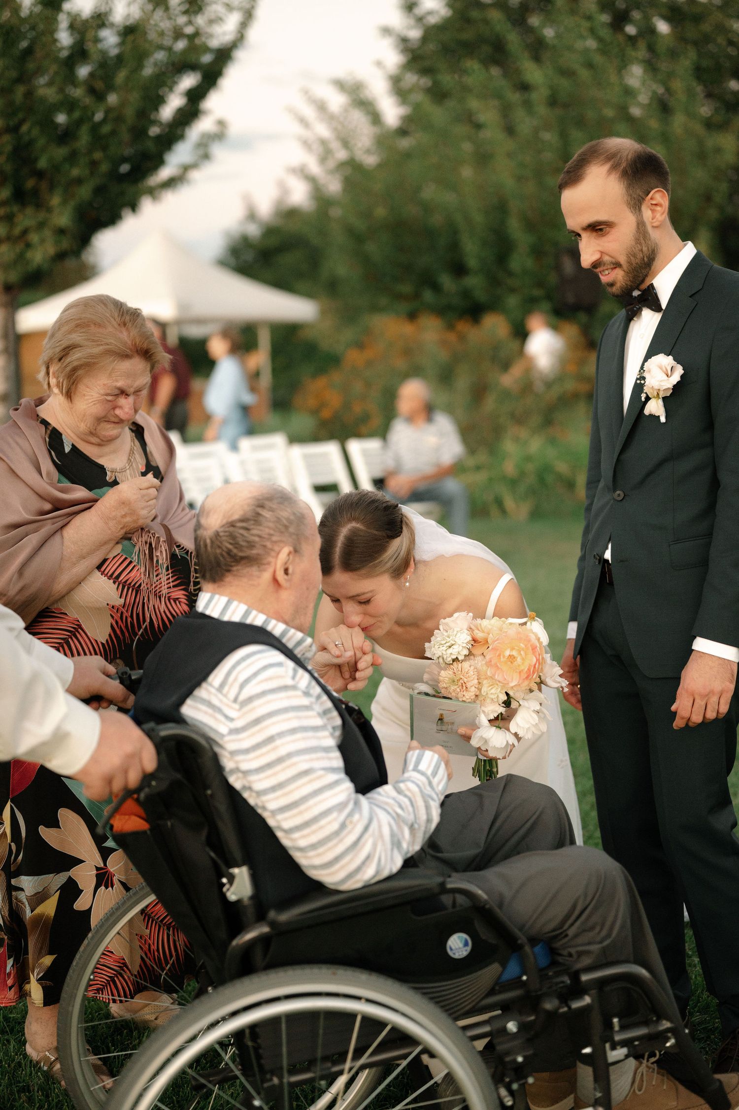 Emotional moment at outdoor wedding ceremony as couple shares special moment with elderly guest in wheelchair.
