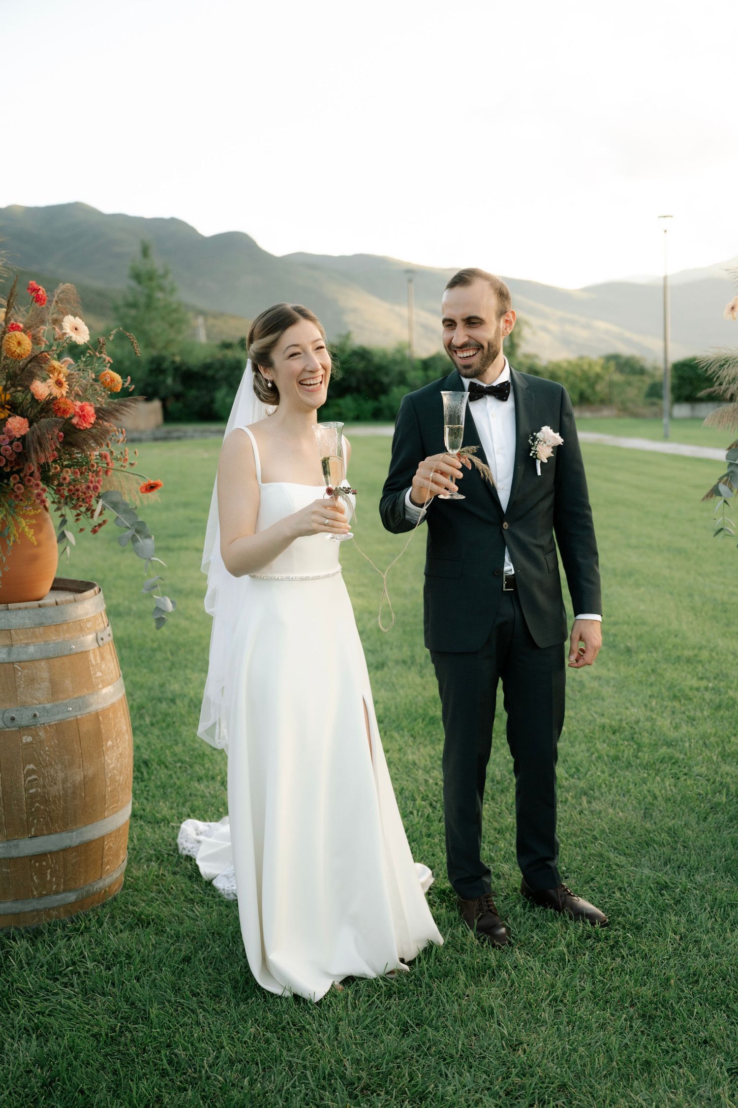 Newlyweds share a joyful moment while holding wine glasses near rustic barrels at an outdoor vineyard wedding reception.