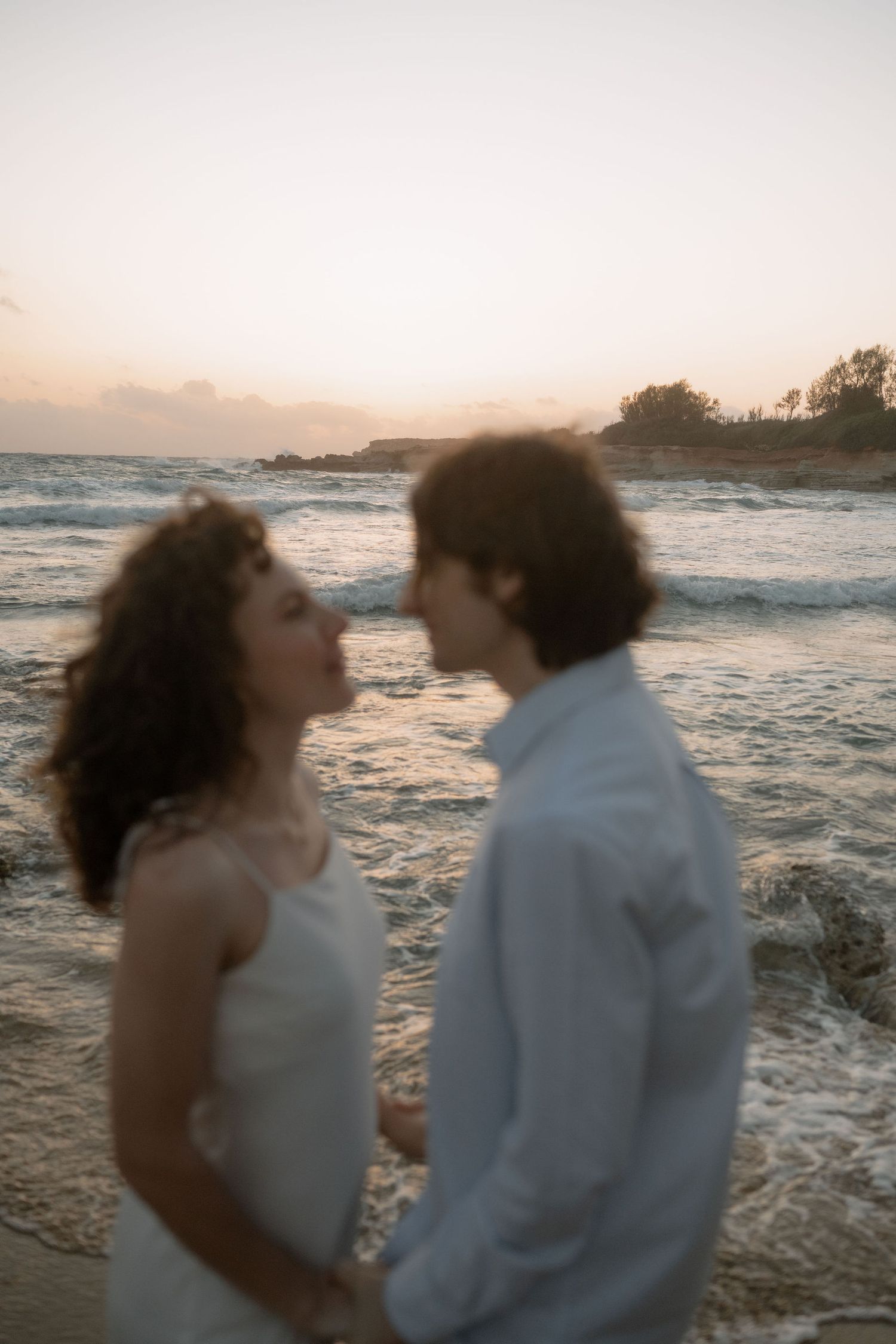 Couple in white attire sharing a romantic moment on a beach at sunset with ocean waves in the background.