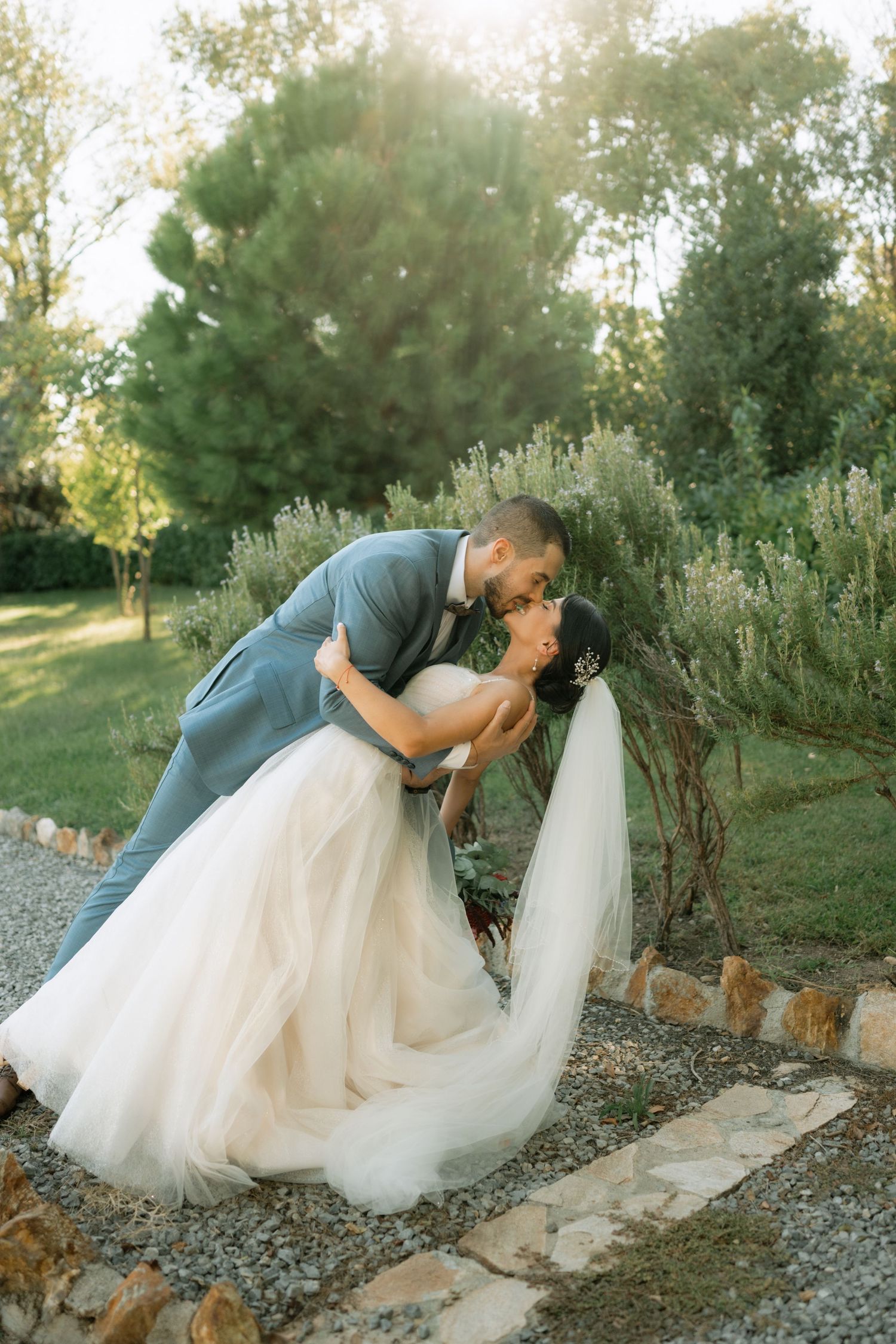 A romantic wedding couple share a kiss in a garden setting with lush greenery and a stone path.