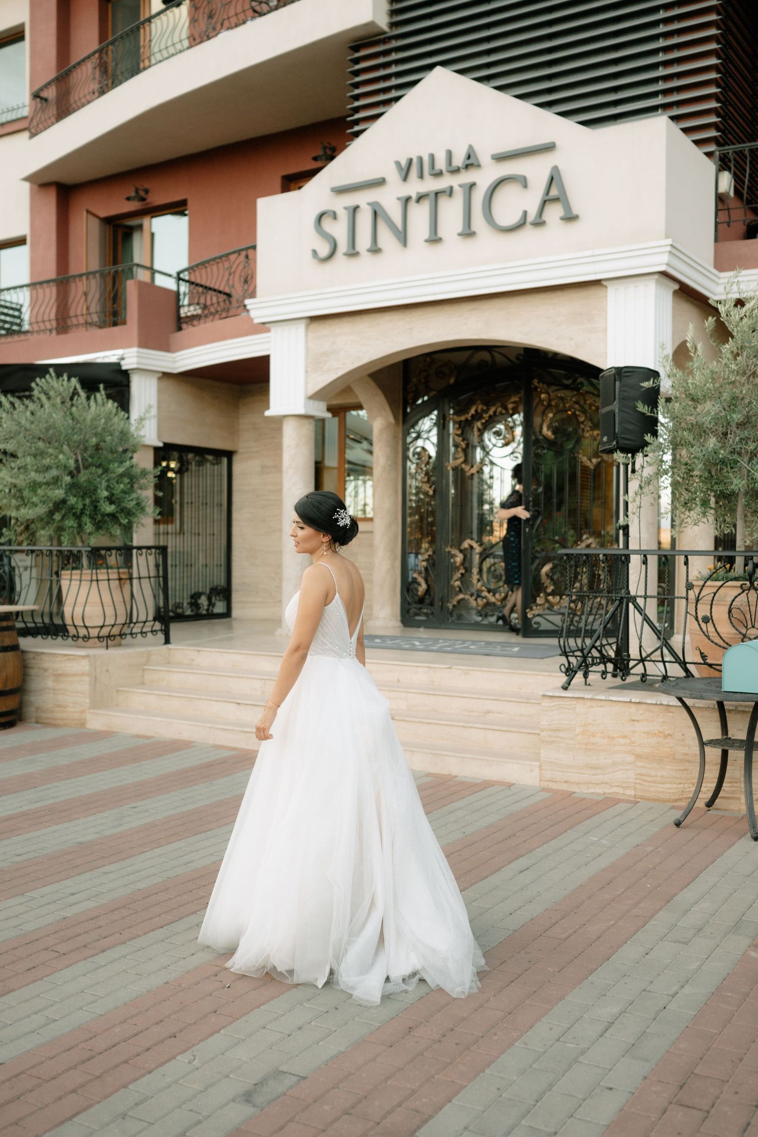 A bride in a flowing white wedding dress twirls gracefully on an outdoor terrace with stone columns and benches.