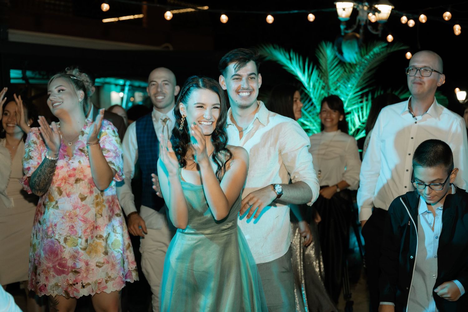 Wedding reception guests dance and celebrate on a dance floor illuminated by warm lights and green uplighting.