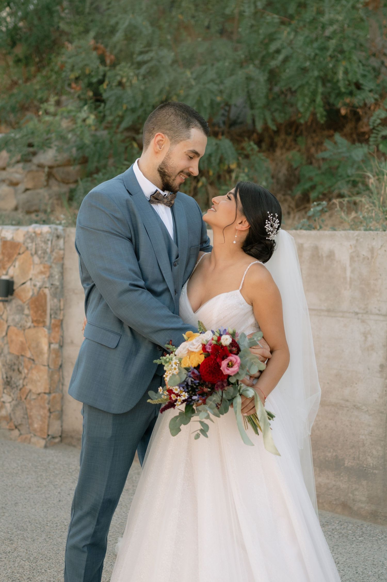 A romantic wedding couple in grey suit and white dress share intimate moments against a stone wall backdrop.