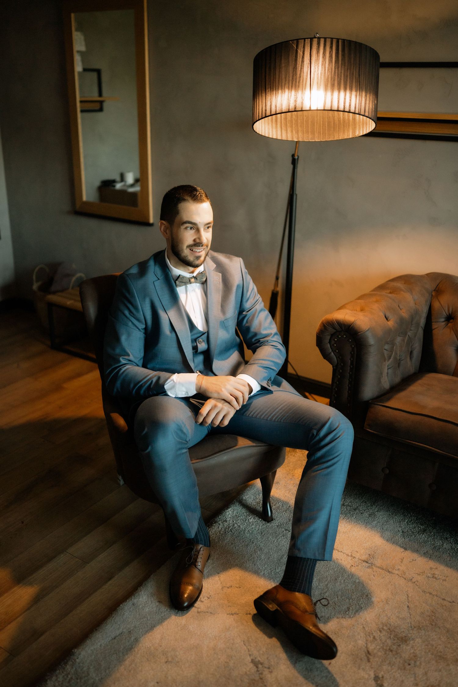 A person in a blue suit sits on a gray couch in a dimly lit room with warm lighting from above.