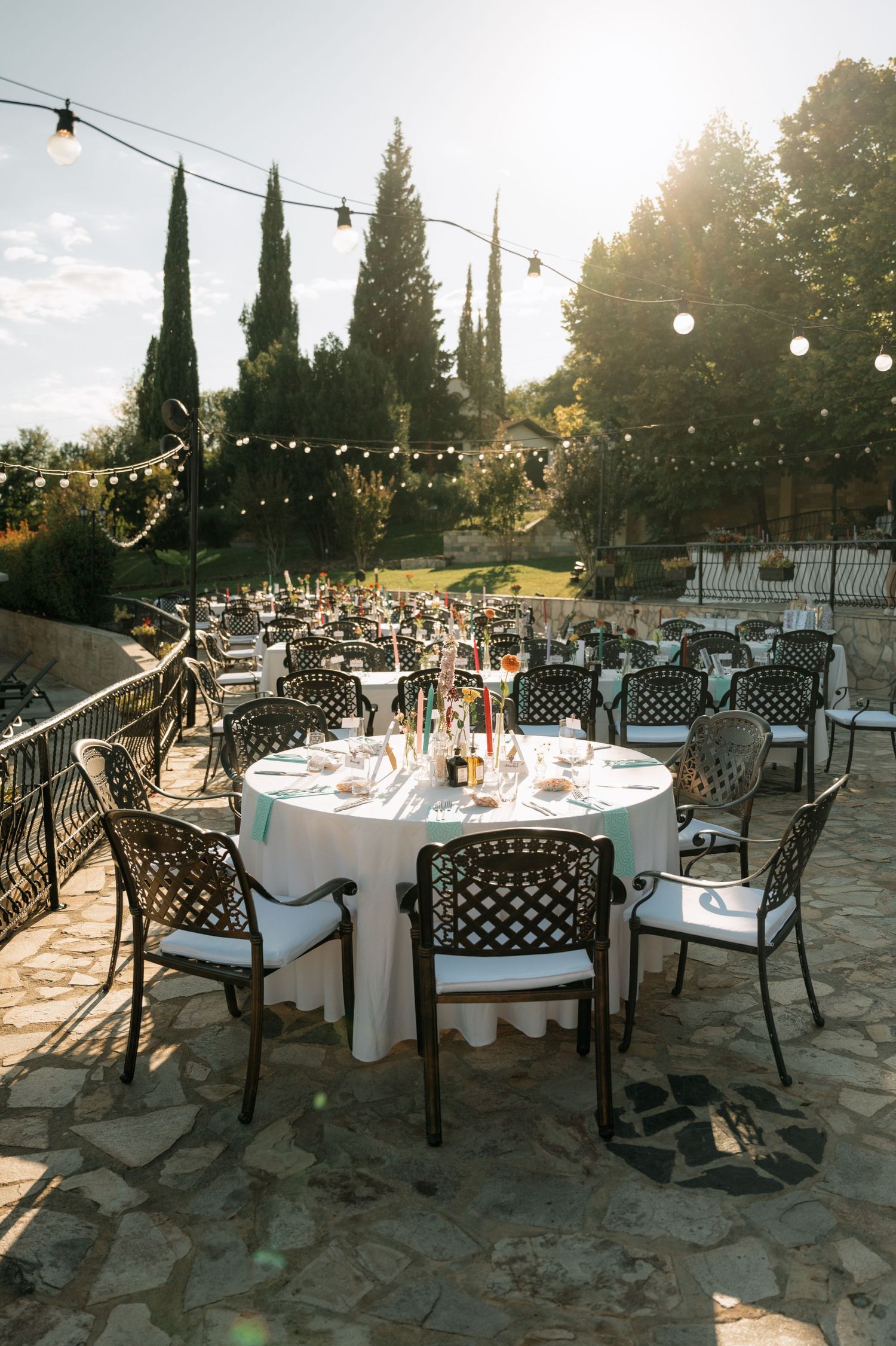 Outdoor wedding reception setup with string lights and cypress trees at sunset on a stone patio.