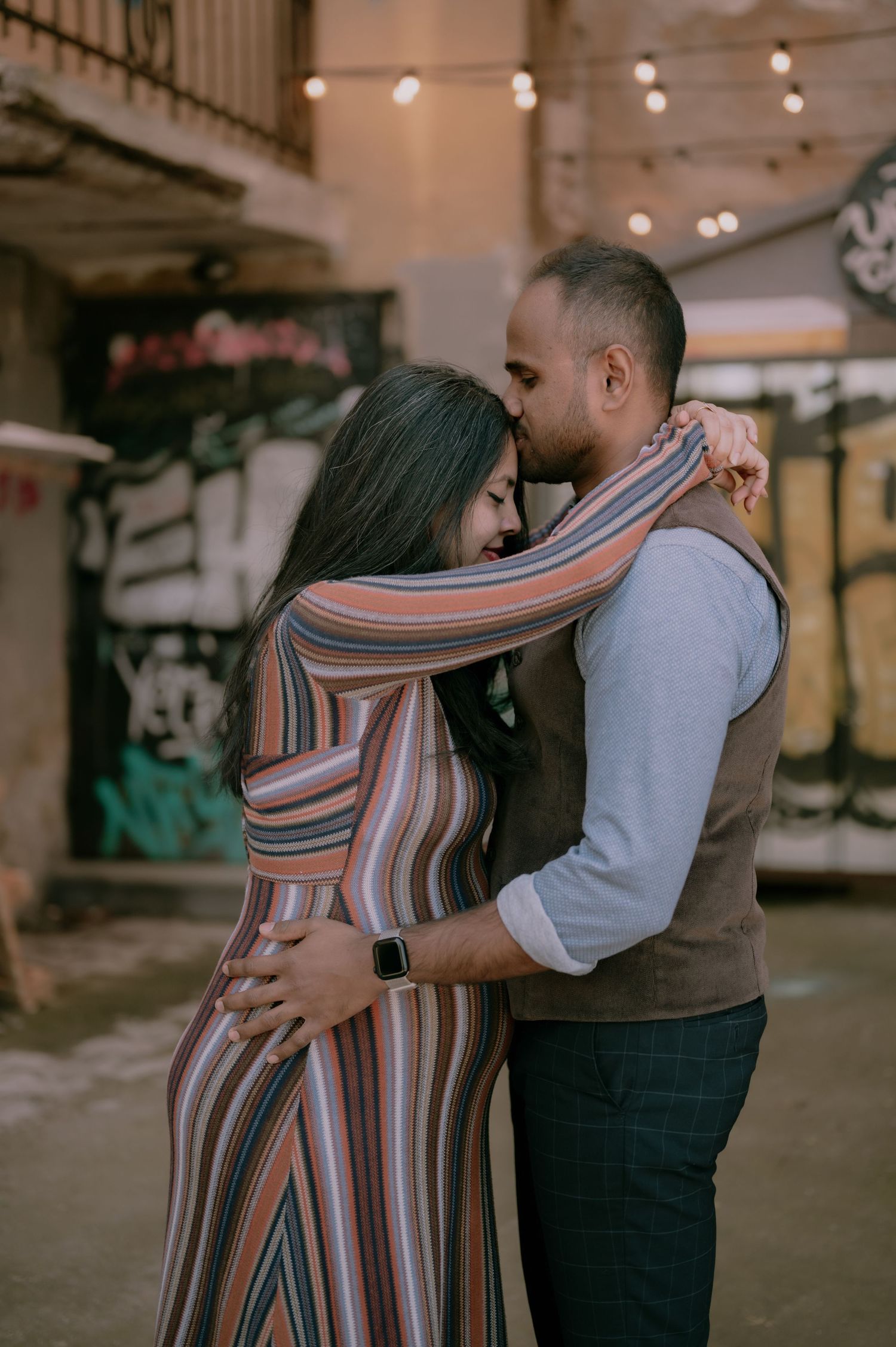 A couple embraces tenderly in an urban alley, woman in colorful striped dress, man in gray shirt and vest.