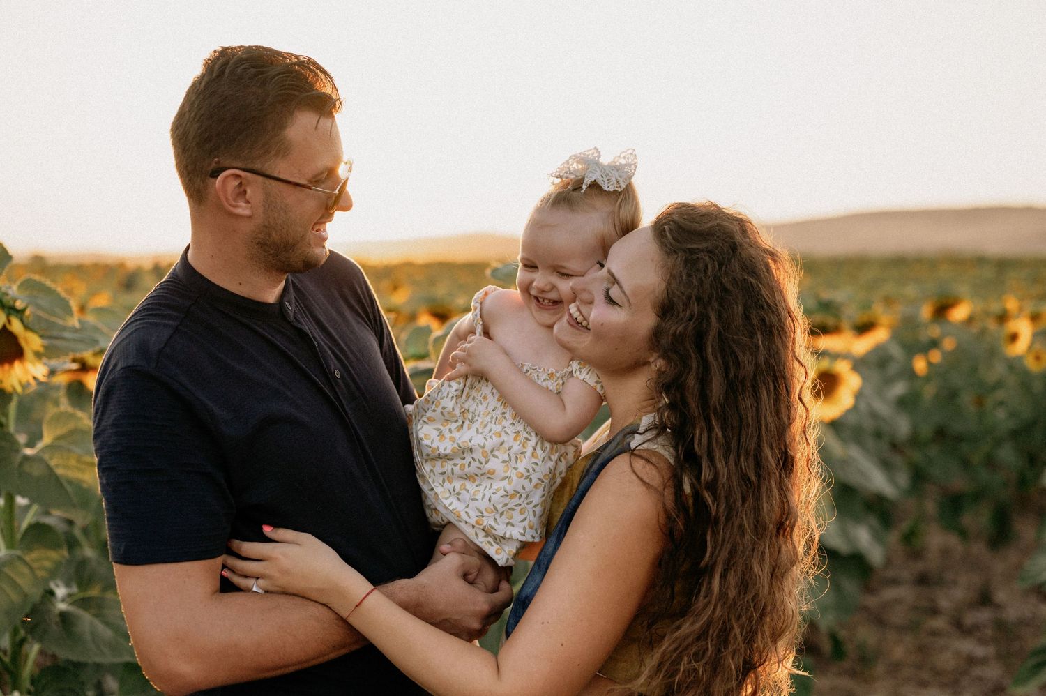 A couple shares a tender moment with their baby girl in a sunflower field at golden hour sunset.