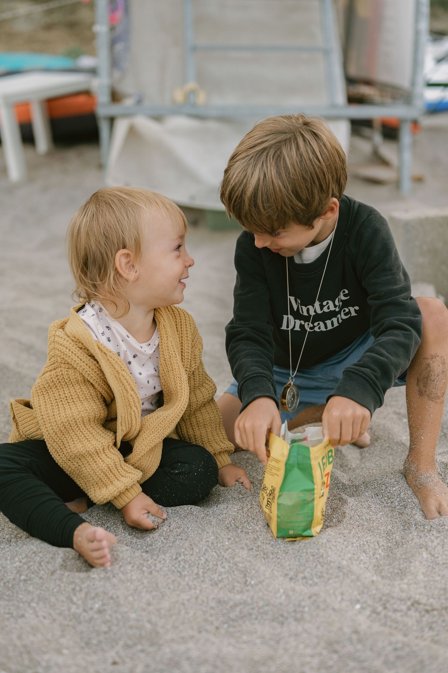 Two young children sit on sandy ground, smiling at each other, one wearing a yellow cardigan and the other a dark sweatshirt.