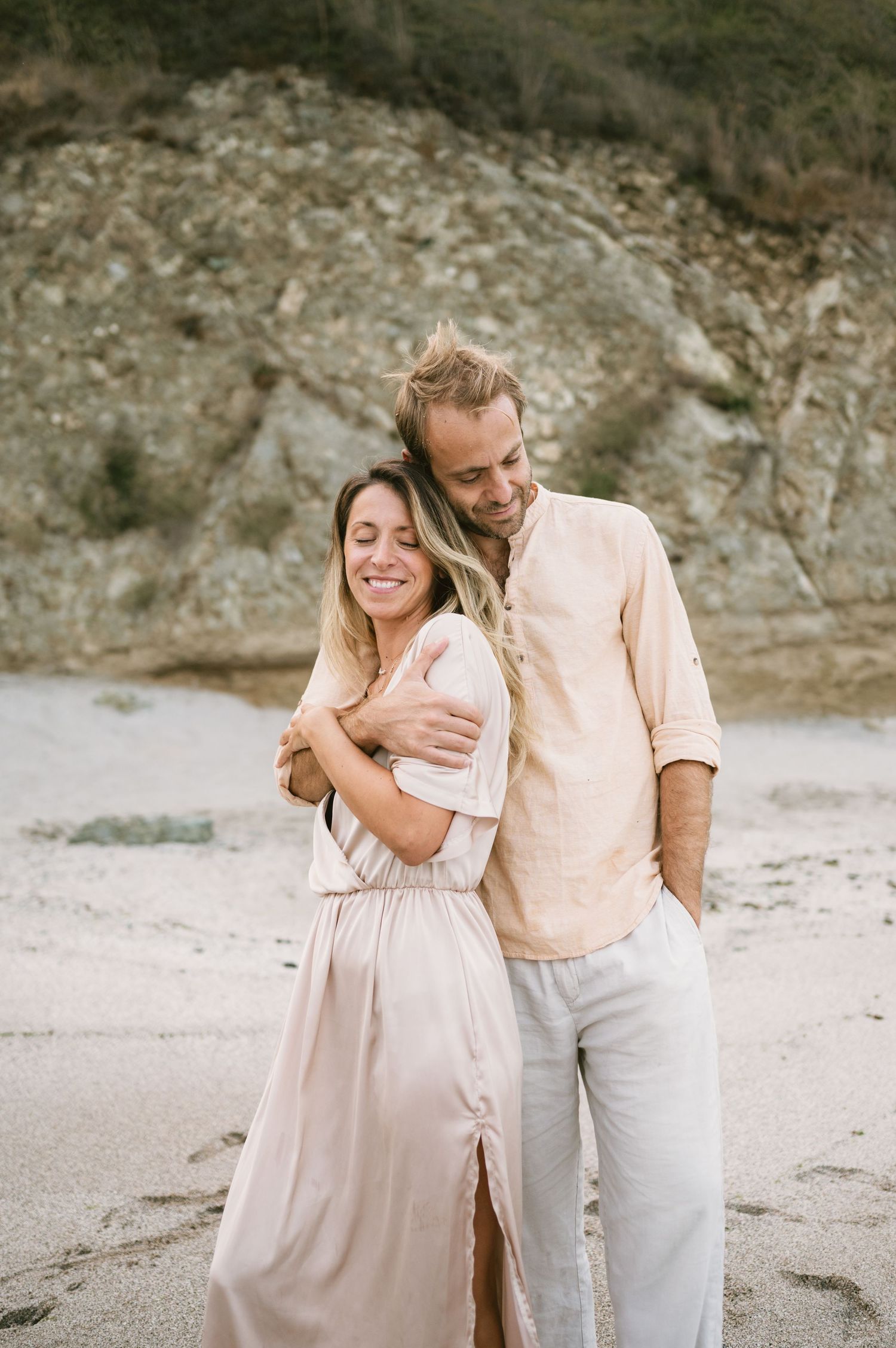 A couple embraces on a sandy beach, smiling warmly, dressed in soft neutral tones with rocky cliffs behind them.