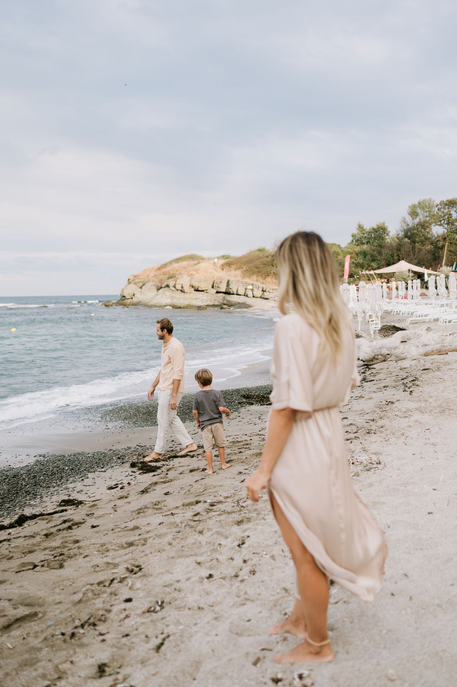Family walking on a sandy beach, woman in pink dress, man and child near ocean waves on a cloudy day.
