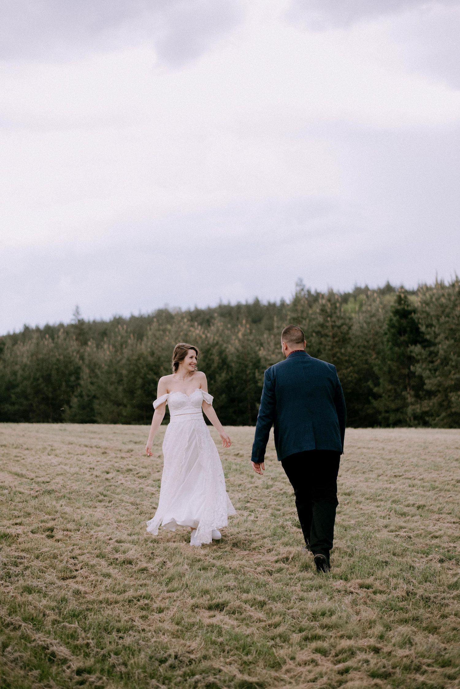 A couple walks up a grassy hill surrounded by pine trees on an overcast day.