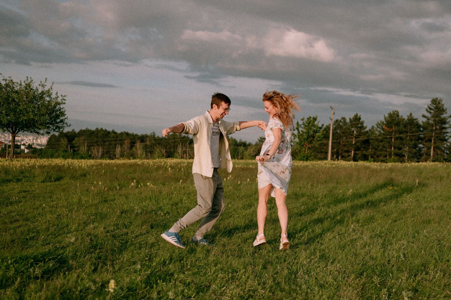 A couple dances and spins joyfully across a green hilltop field at sunset.