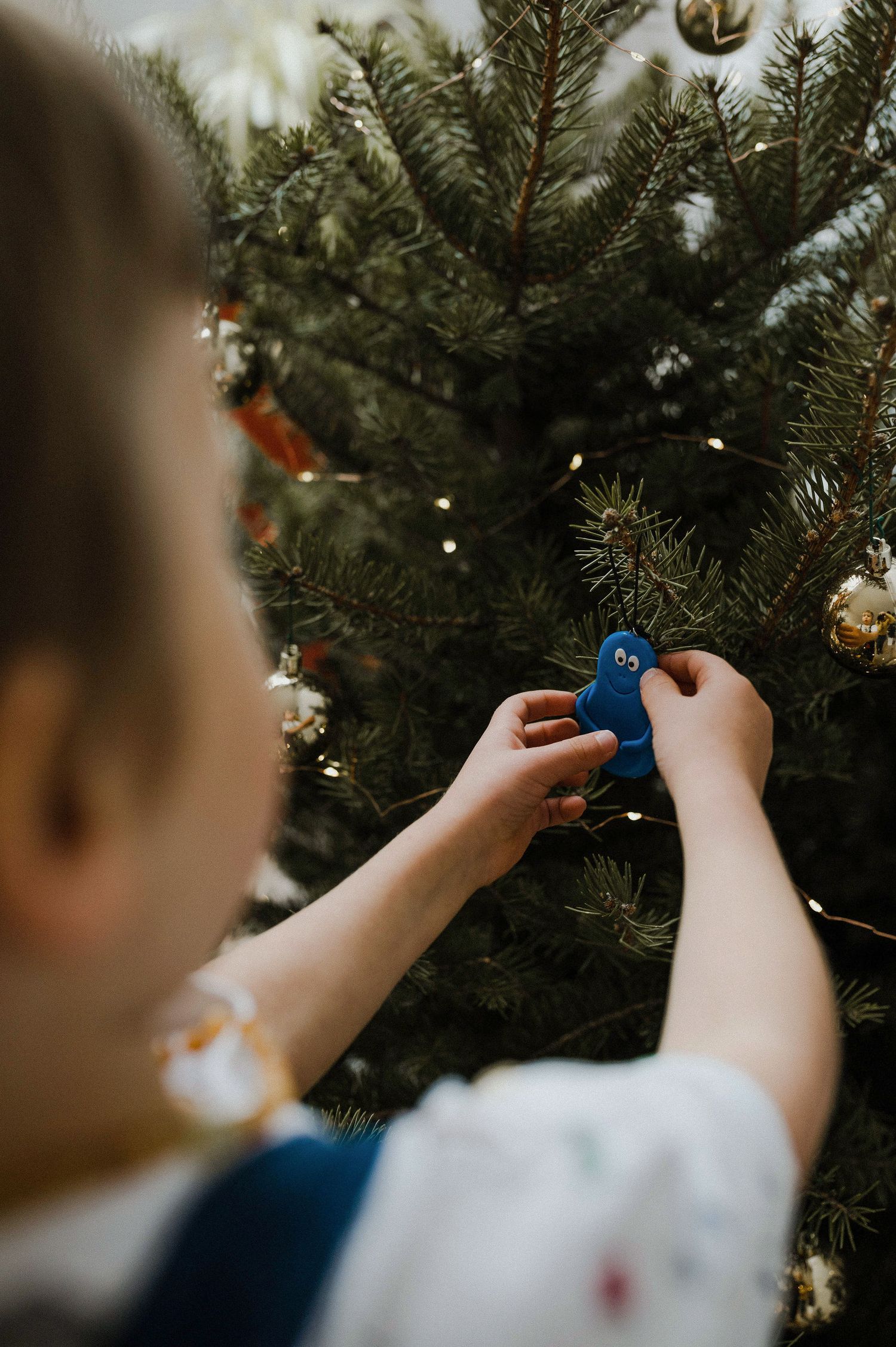 Small hands carefully place a blue ornament on an evergreen Christmas tree decorated with twinkling lights.