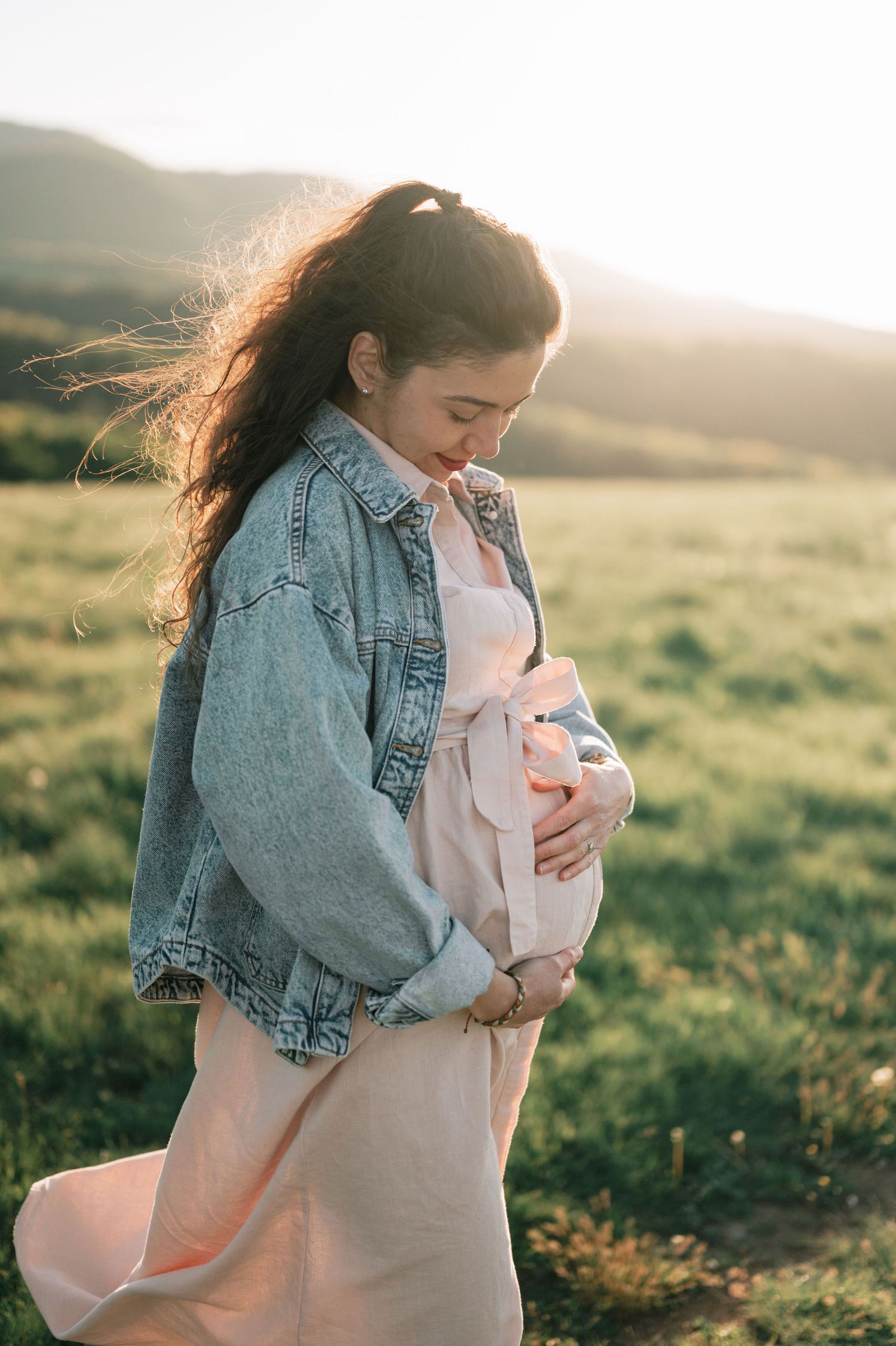 A person wearing a denim jacket holds a baby in a peaceful sunlit field during golden hour.