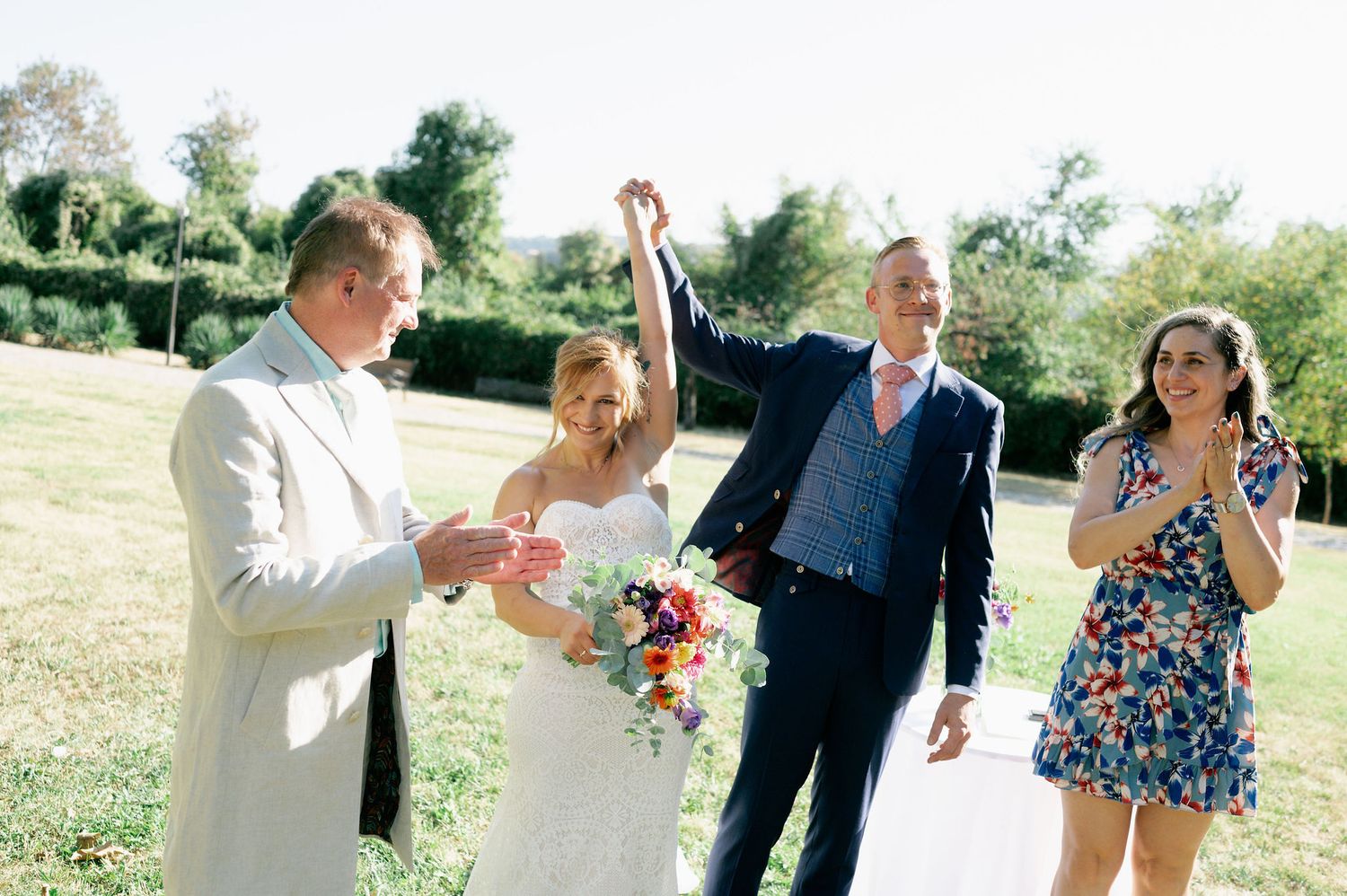 Wedding party members celebrate with joyful expressions in a sunny outdoor setting.