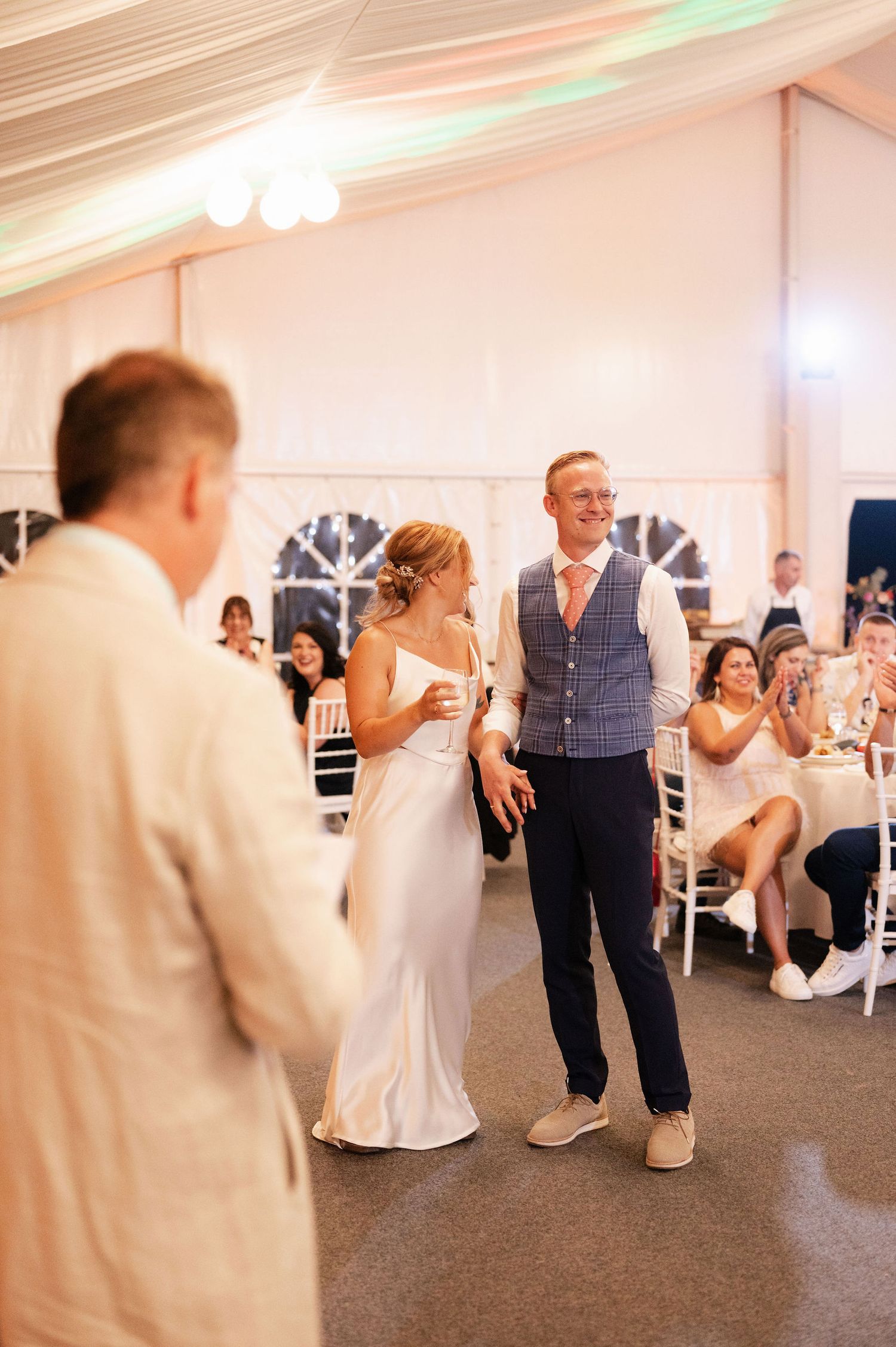 Wedding reception guests enjoying speeches in a beautifully decorated tent venue with white chairs.