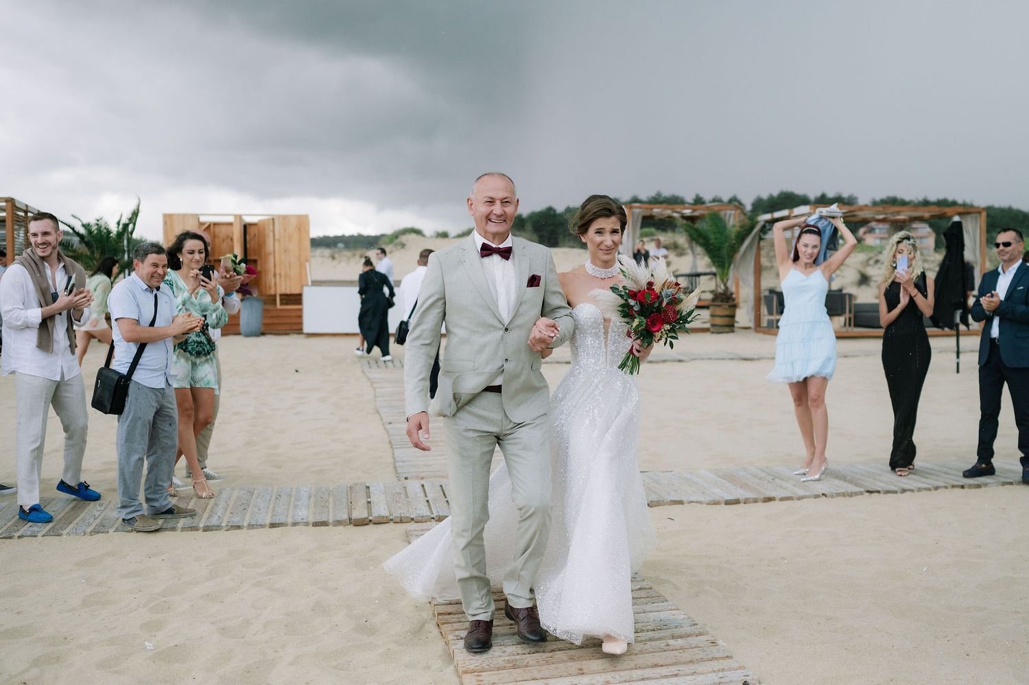 A couple in wedding attire walks together on a sandy beach boardwalk surrounded by guests.