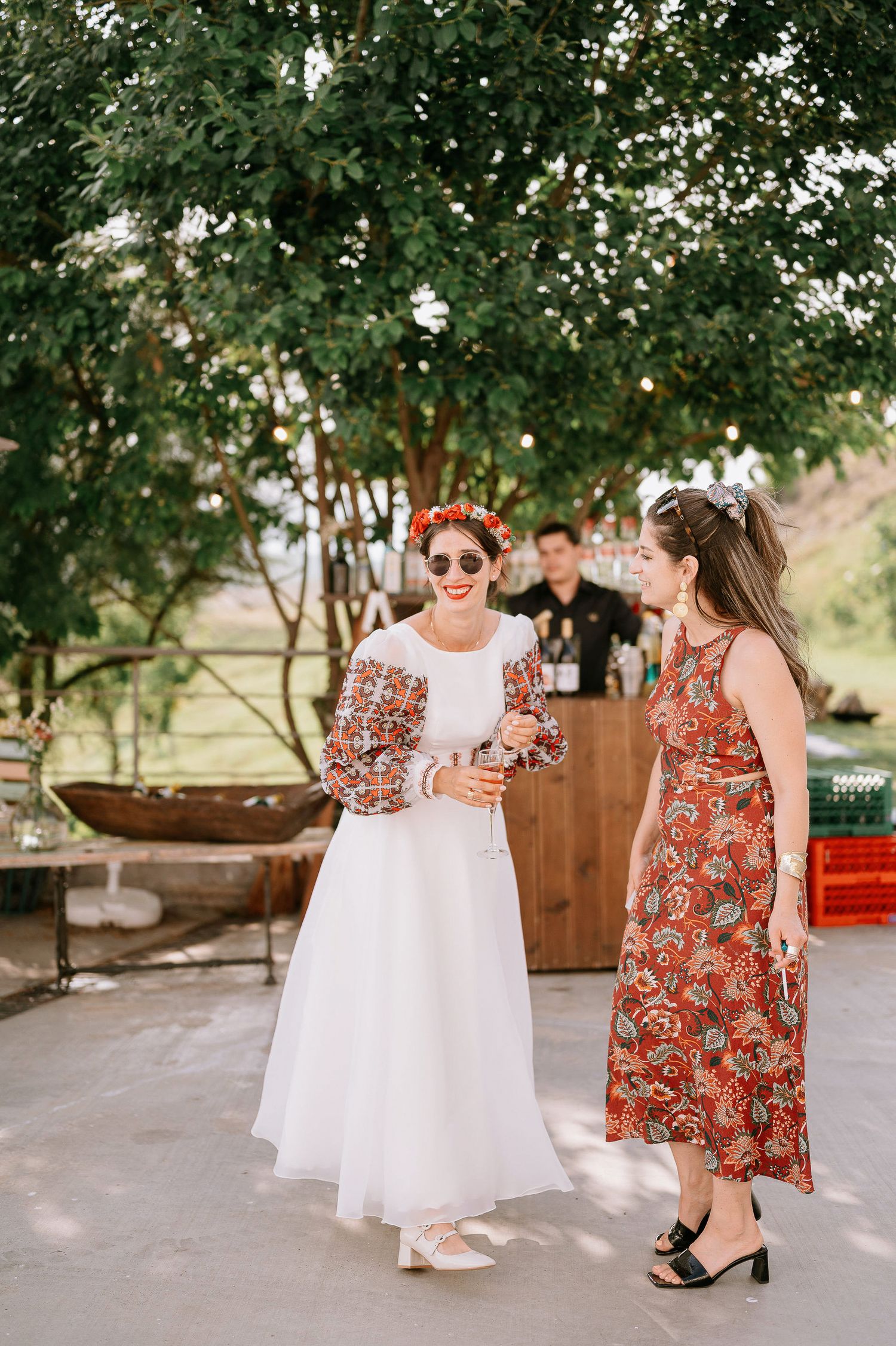 Two women in floral dresses chat during an outdoor wedding reception.
