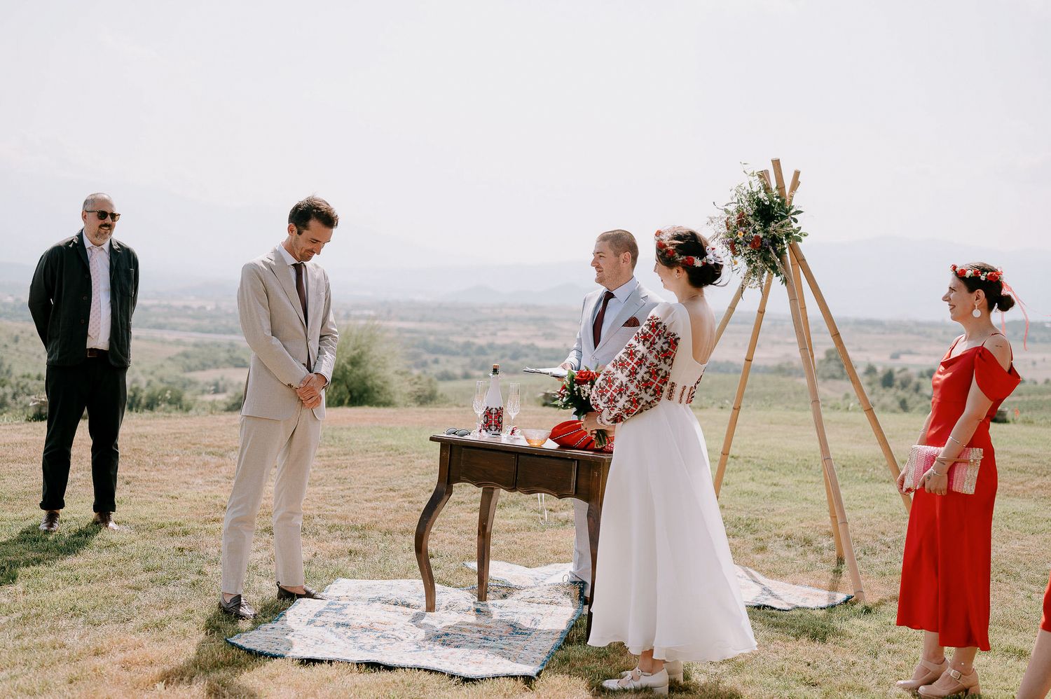 Wedding guests and wedding party gather around a rustic wooden arch on a hilltop for an outdoor ceremony.