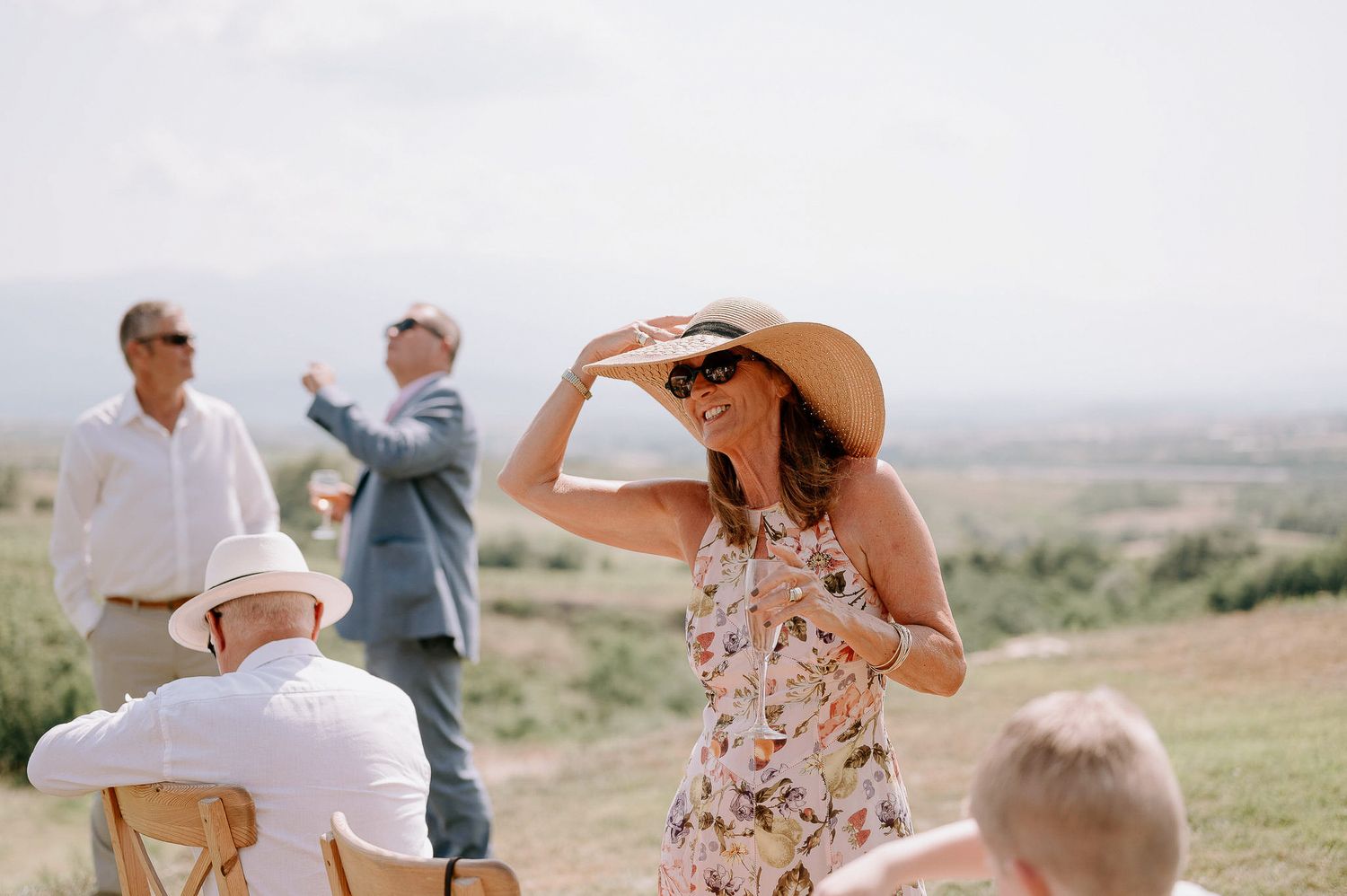 Summer wedding guests in casual attire gather on scenic hilltop overlooking valley.