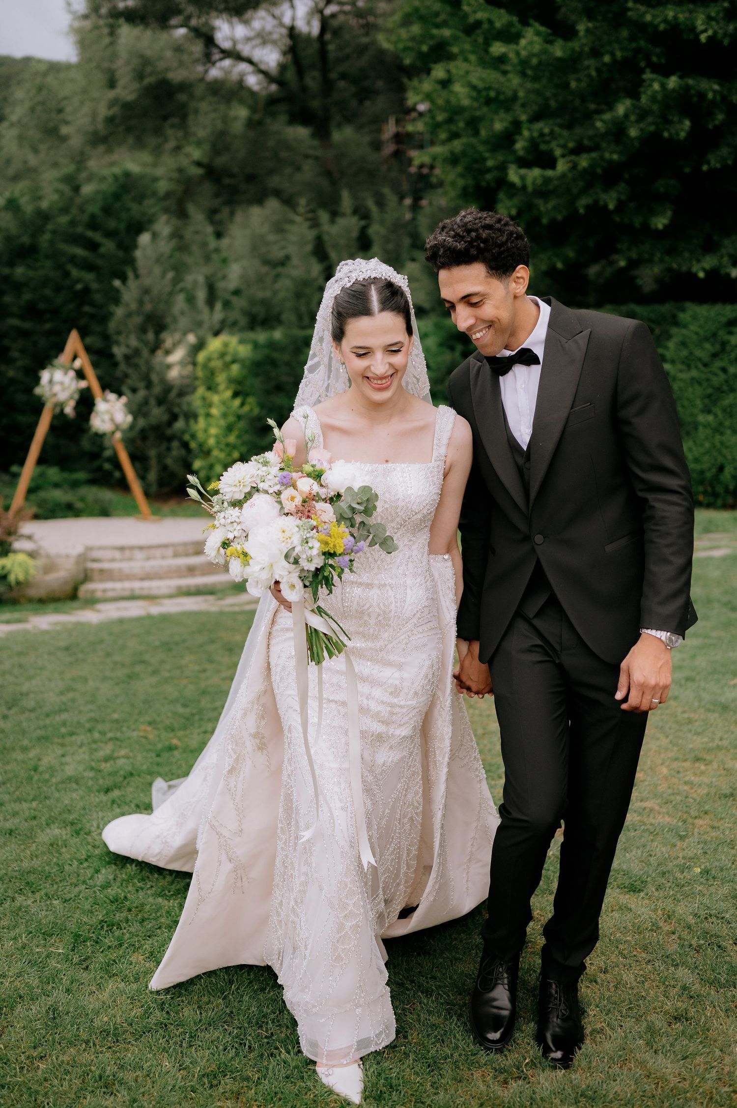 A couple walking together on grass, with the bride holding a delicate floral bouquet.