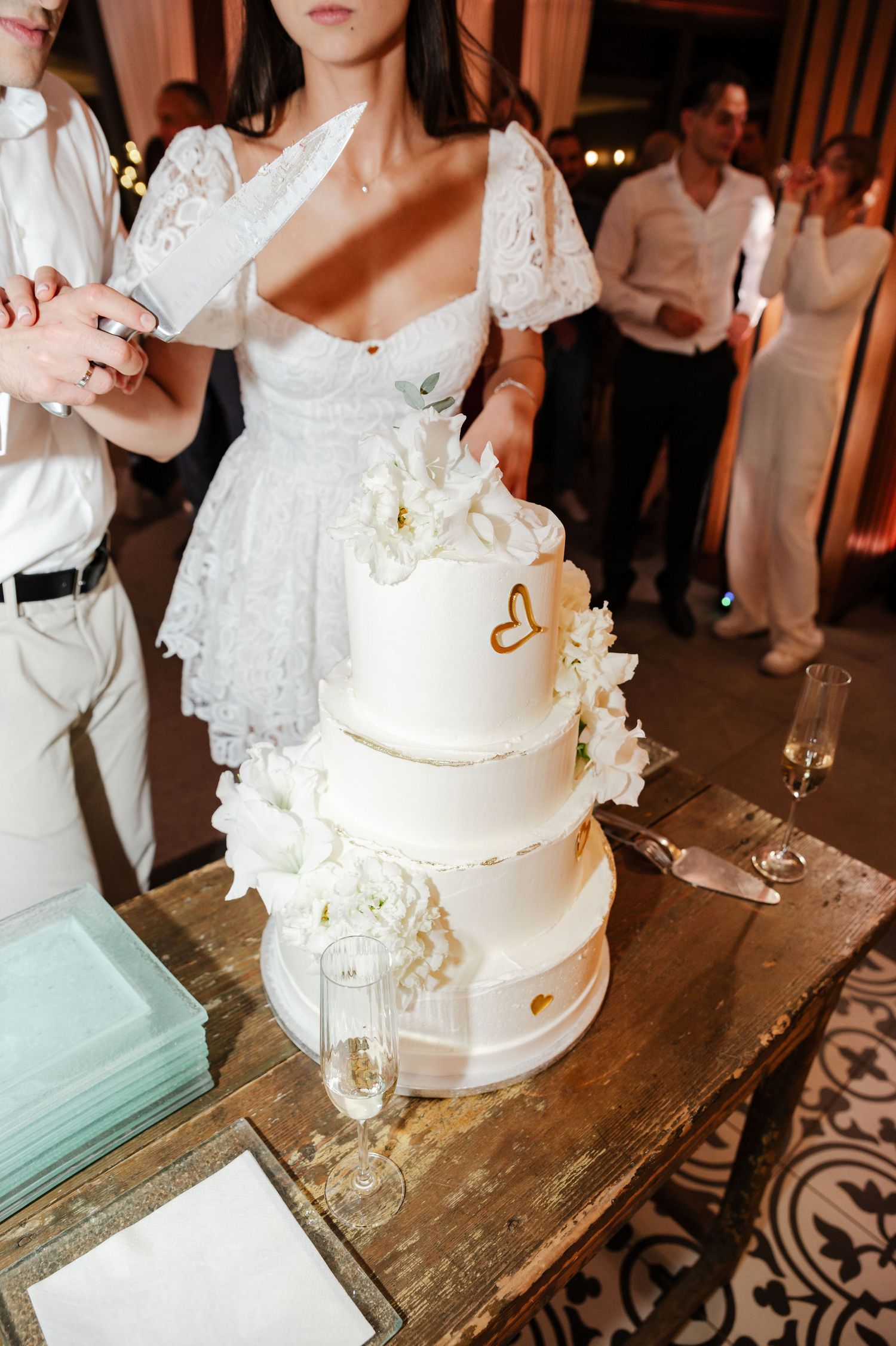 White tiered wedding cake with gold accents displayed on vintage wooden table during reception.