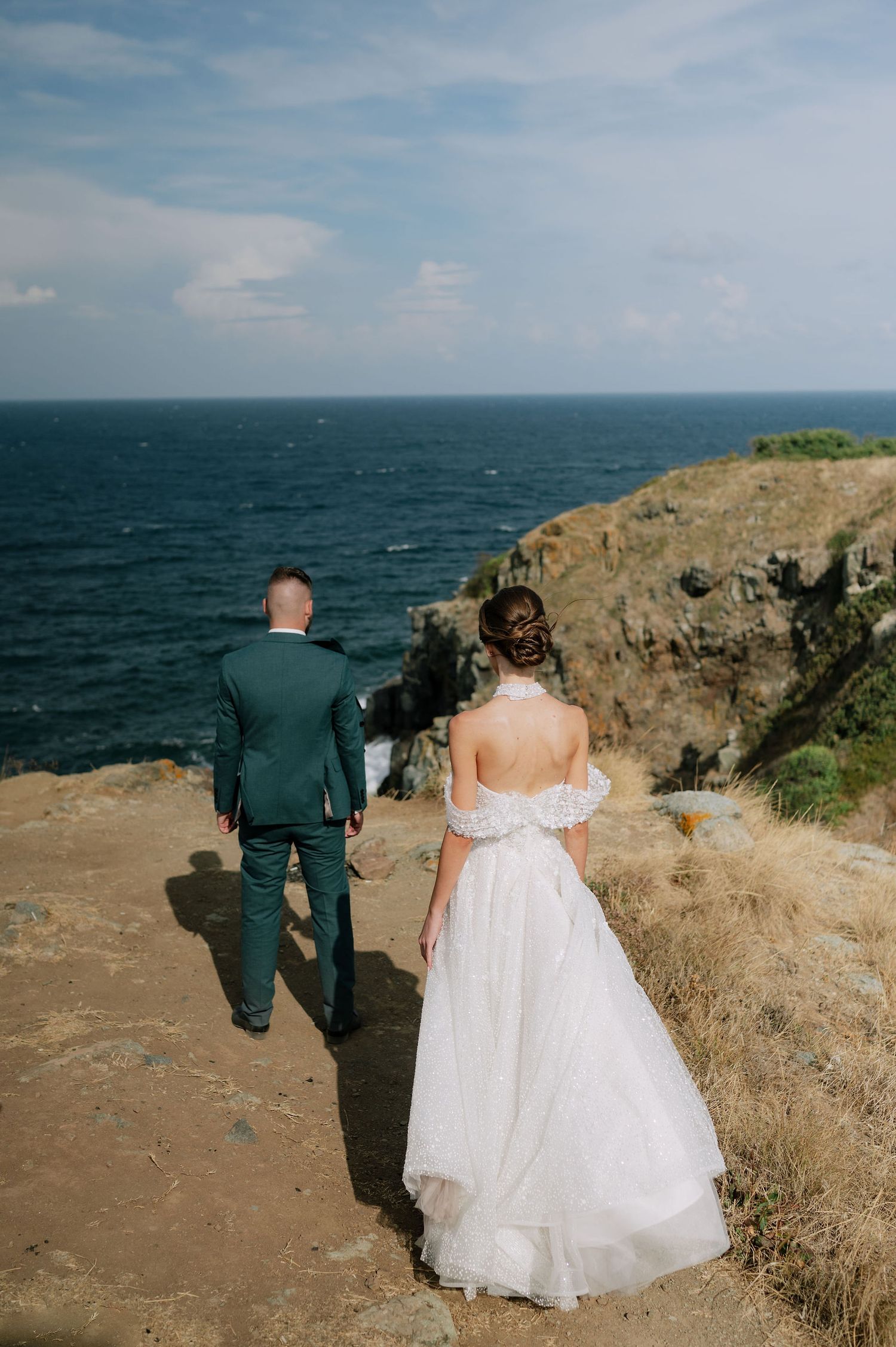 A couple in wedding attire walking along a dramatic coastal cliff edge.